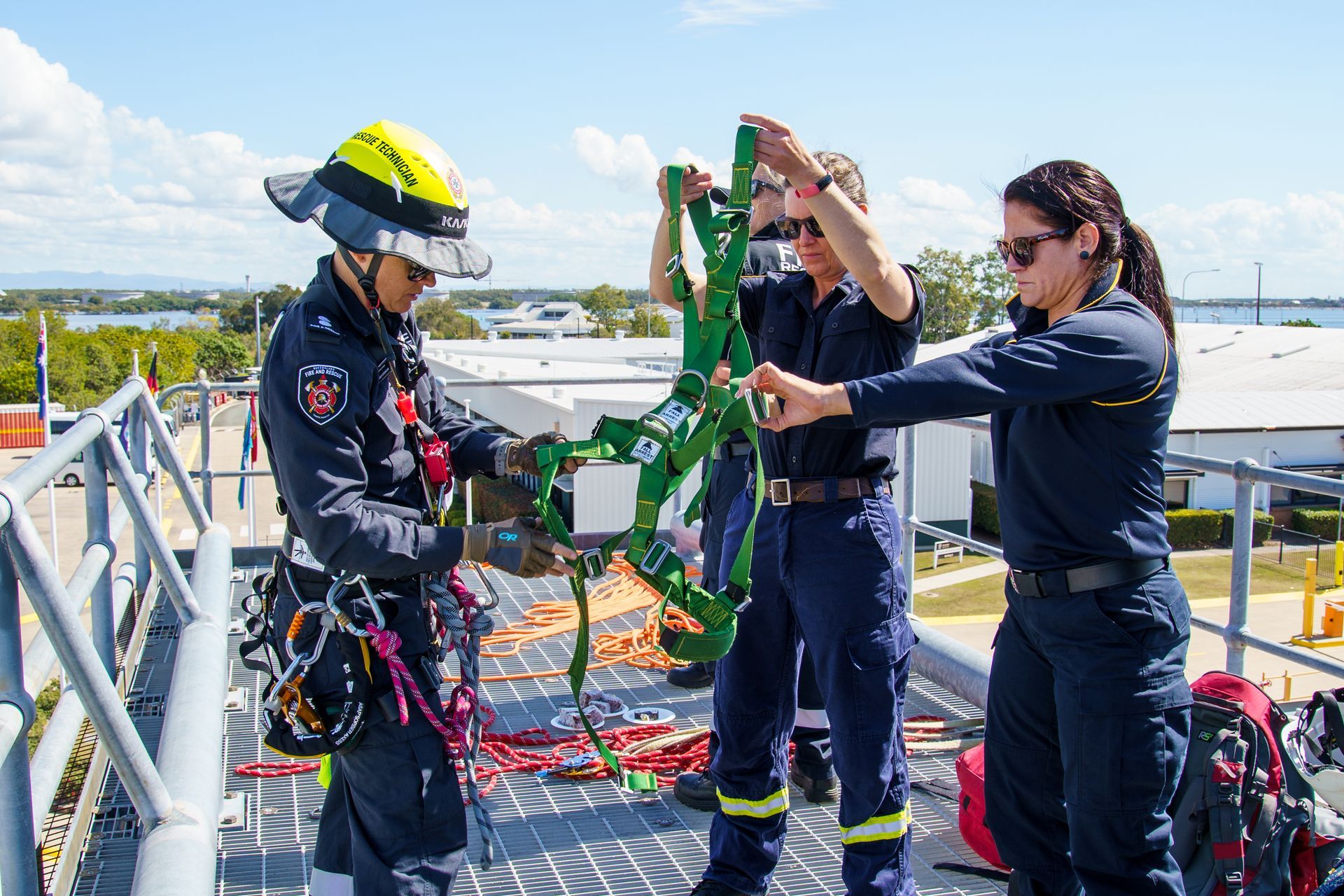 Firefighters on a rooftop training with safety harnesses; one person is wearing a helmet and holding equipment.