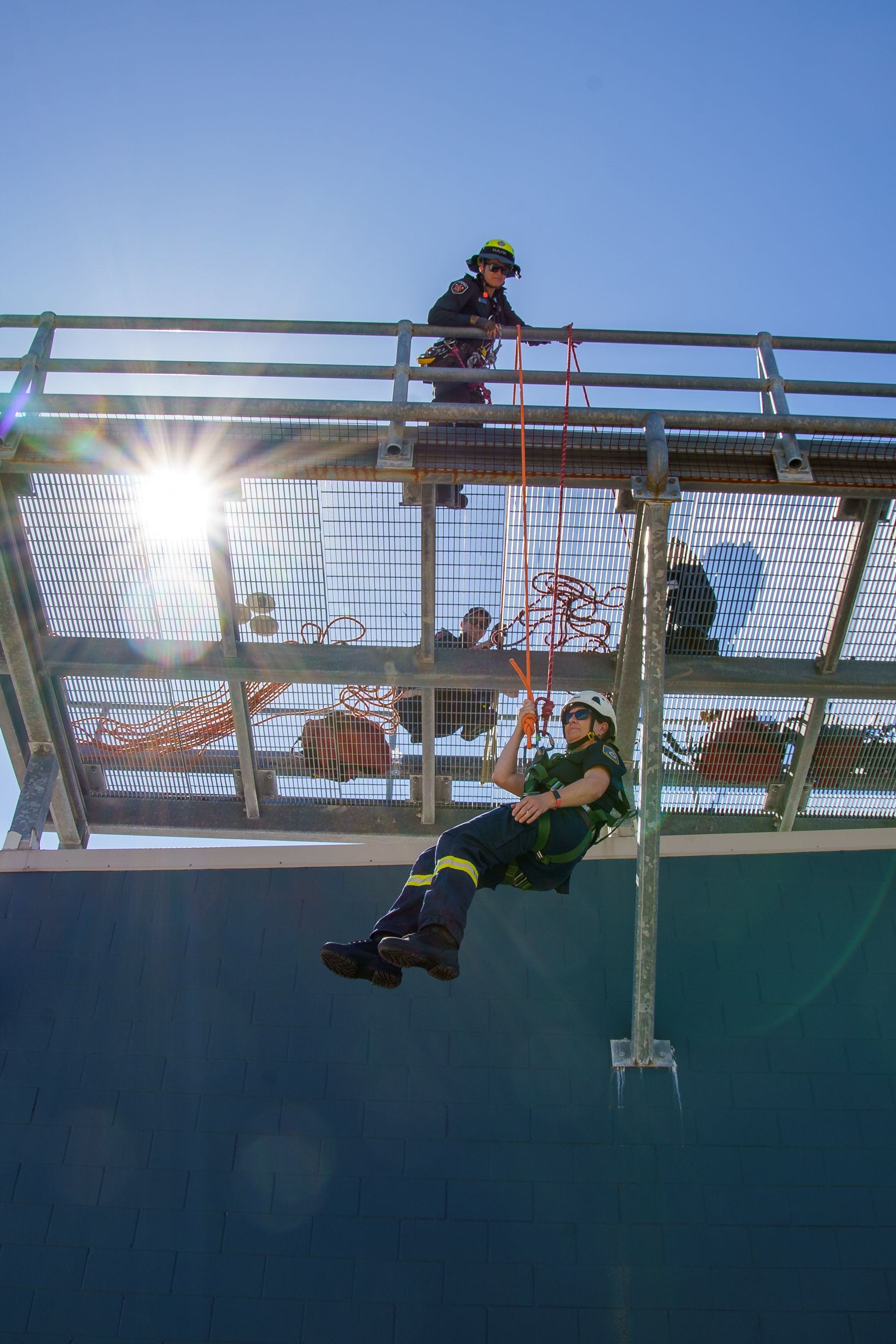 Firefighter rappelling down a blue wall from a metal platform; another firefighter secures the rope on top.