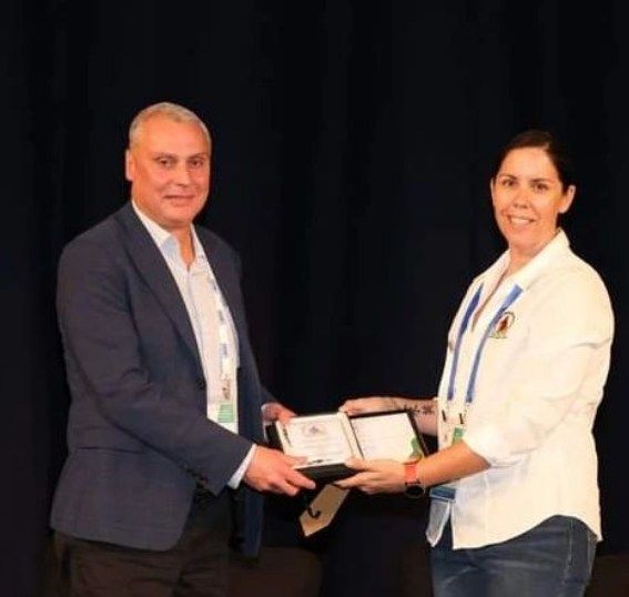 Man in suit presents award to woman in white shirt. They smile, indoors, dark backdrop.