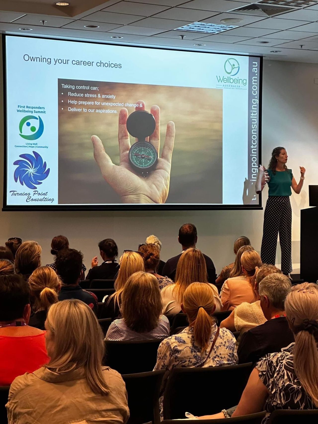 Woman presenting to a crowd, slide with compass graphic. Audience is seated.