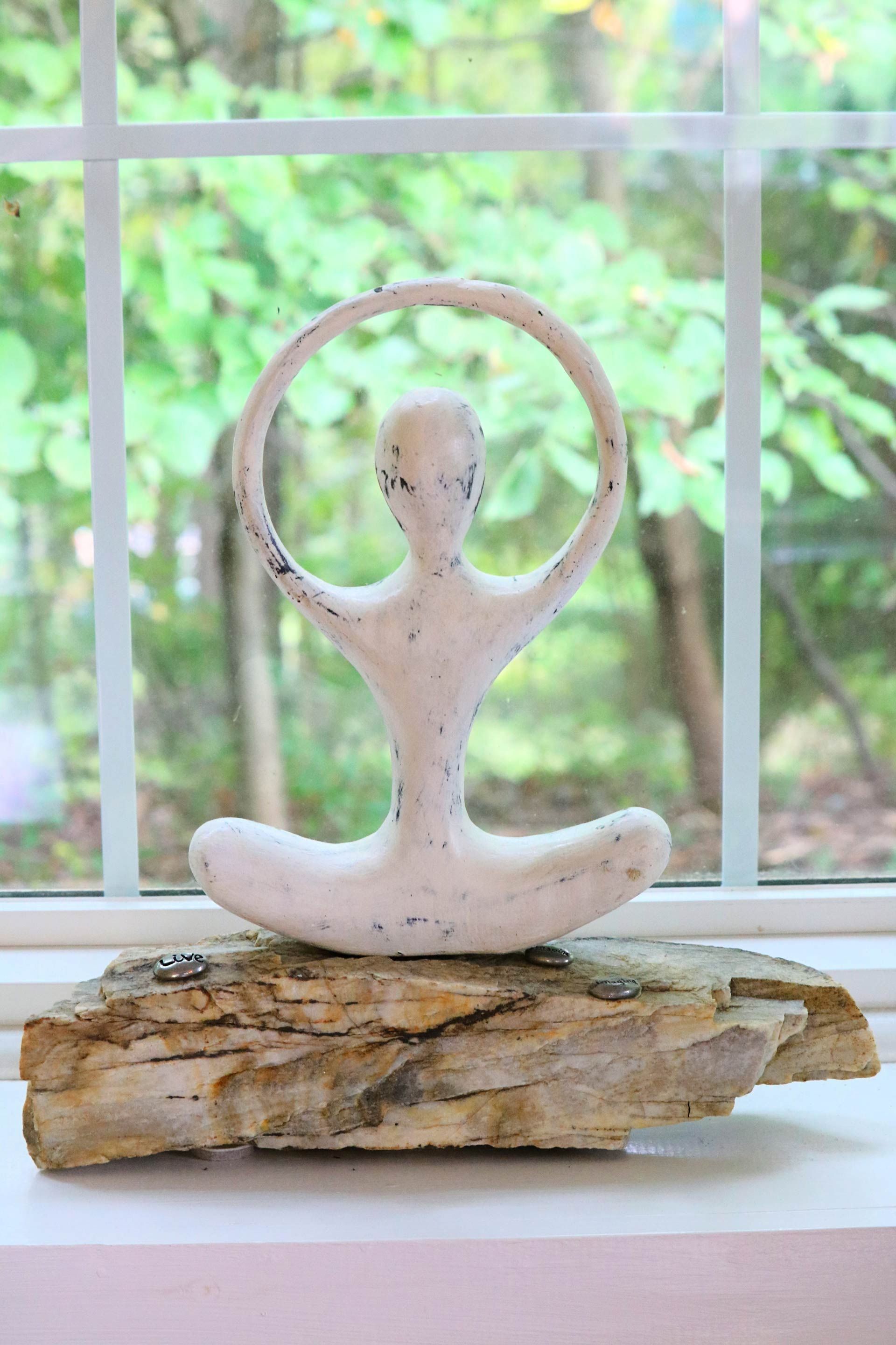 a statue of a woman in a lotus position is sitting on a piece of driftwood in front of a window .