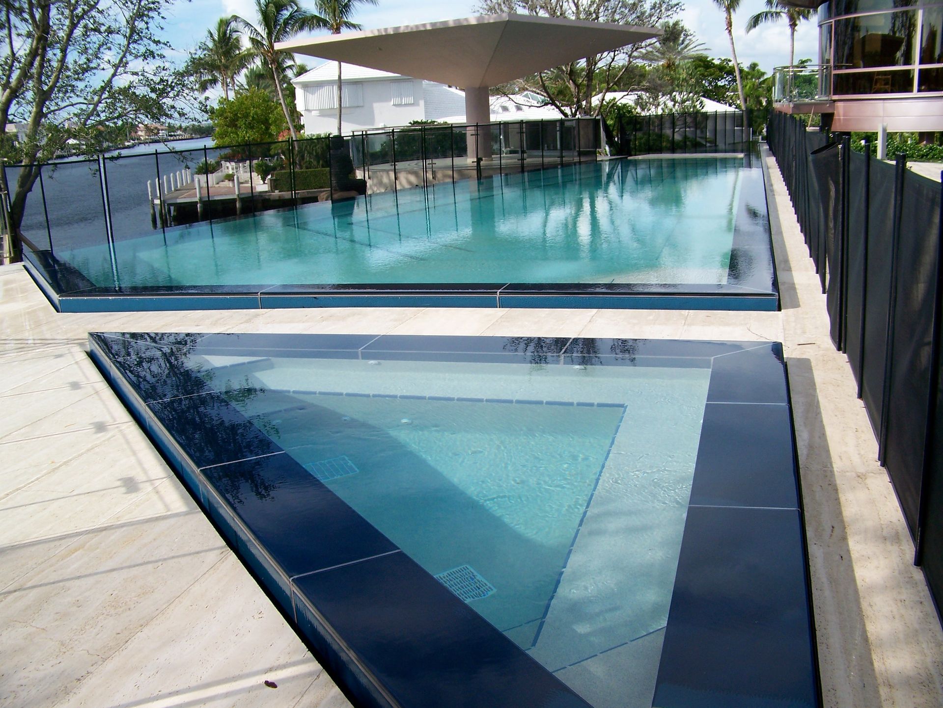 Two-tiered modern pool with glass walls, overlooking a tropical landscape and building. Clear turquoise water.