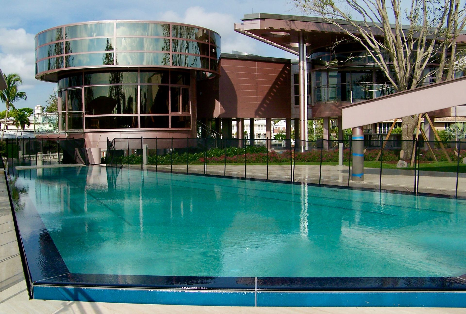 A turquoise pool sits in front of a modern, multi-story building featuring a large glass-walled circular room.