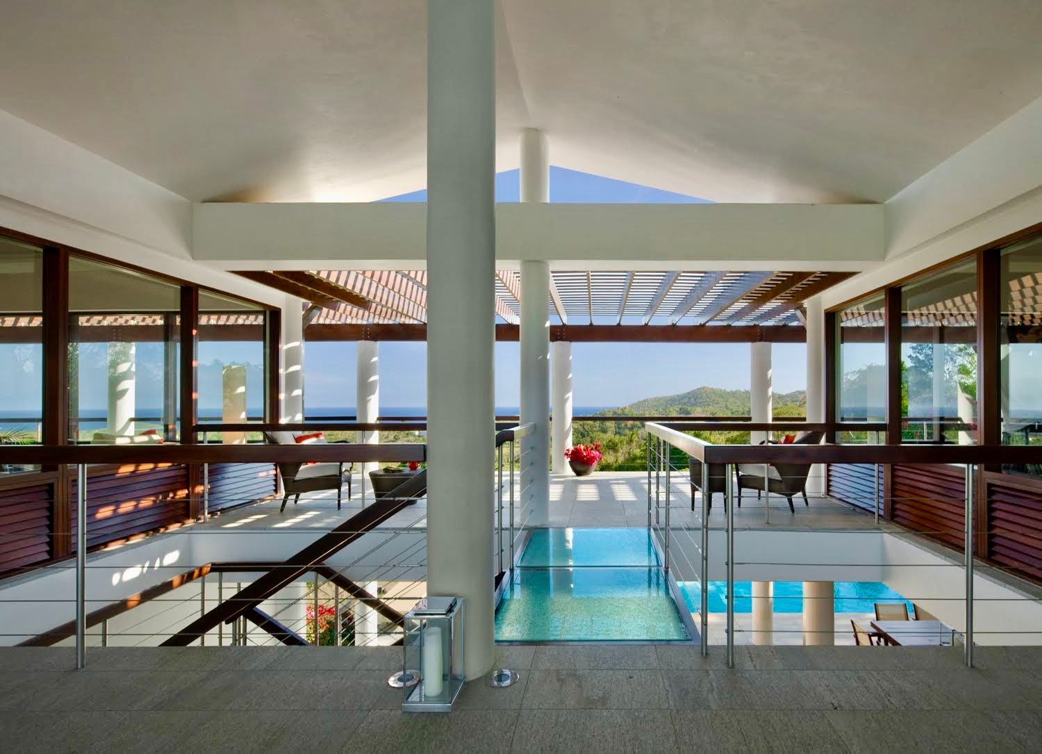 An open-concept interior with a glass walkway overlooking a swimming pool, leading to a scenic tropical outdoor view.