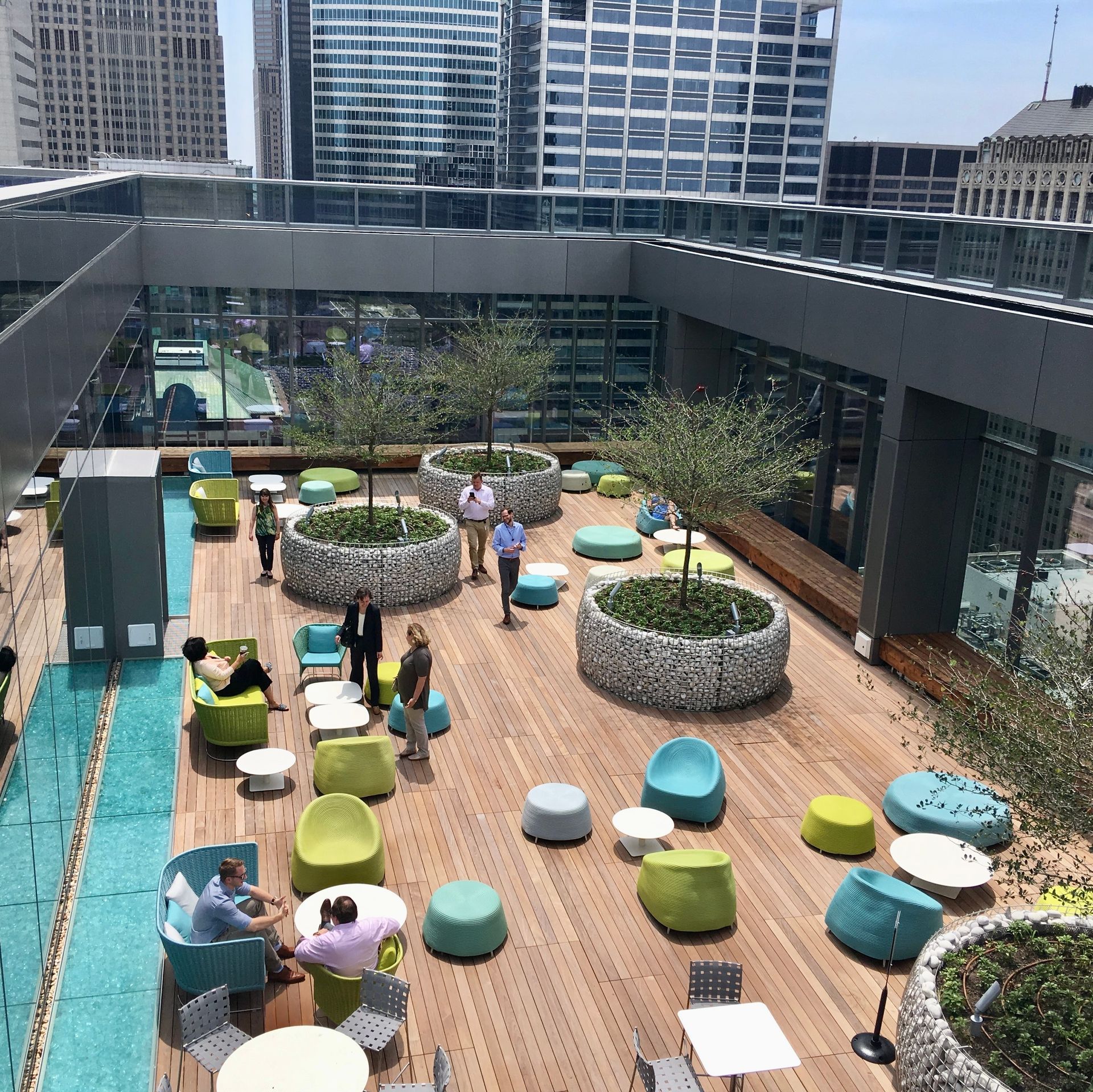 Rooftop patio with trees, seating, and people. Wooden deck, water feature, and cityscape backdrop.