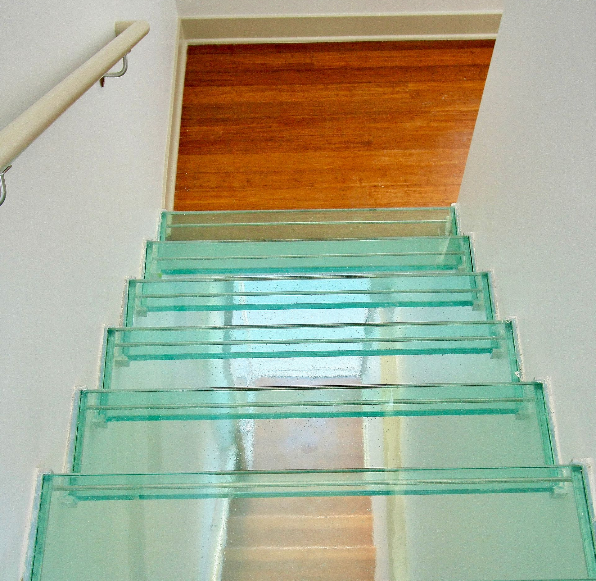 Glass staircase, leading upwards. Green glass steps, wooden floor at the top. White walls.
