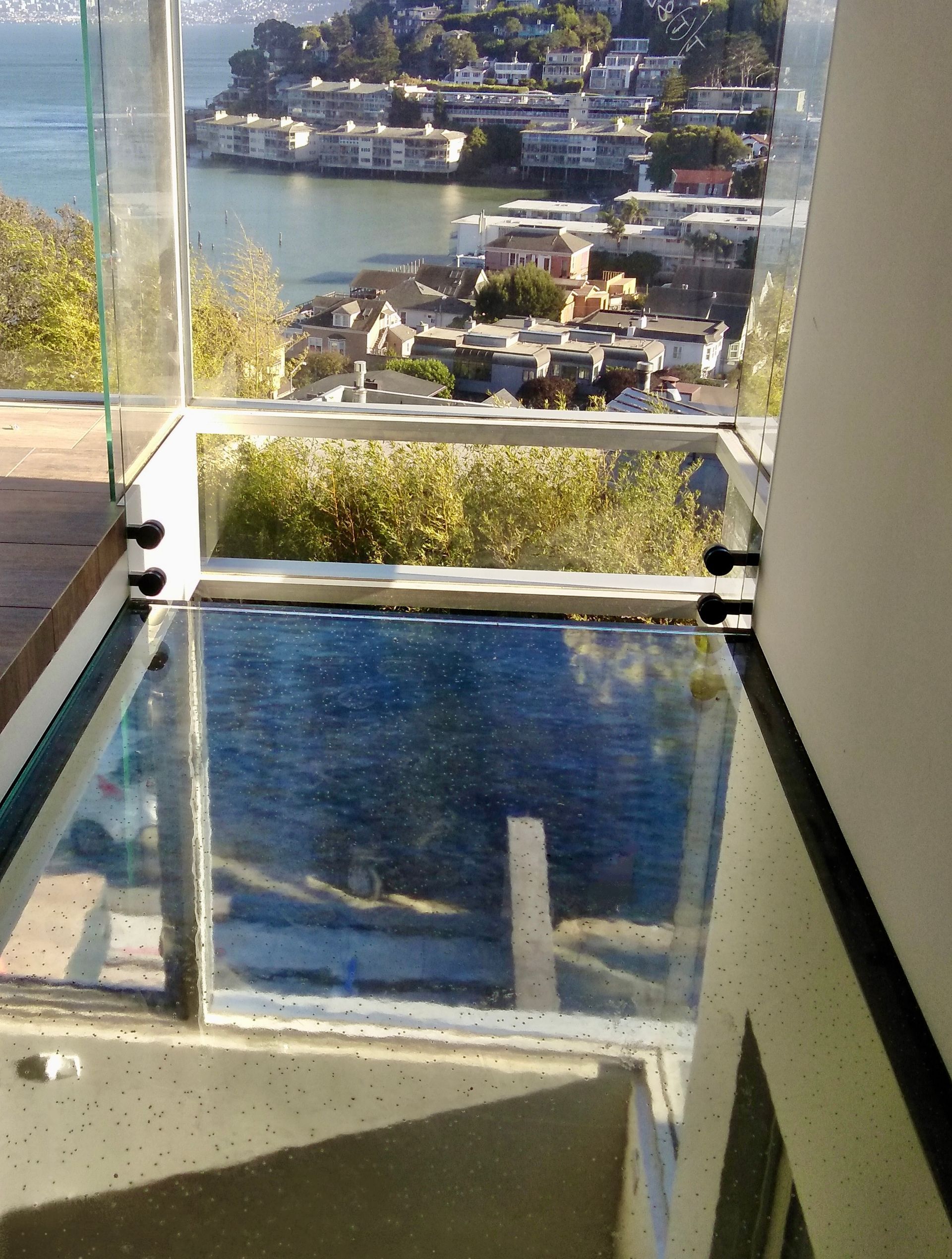 A glass floor balcony overlooking a scenic bay with hillside homes and water visible below.