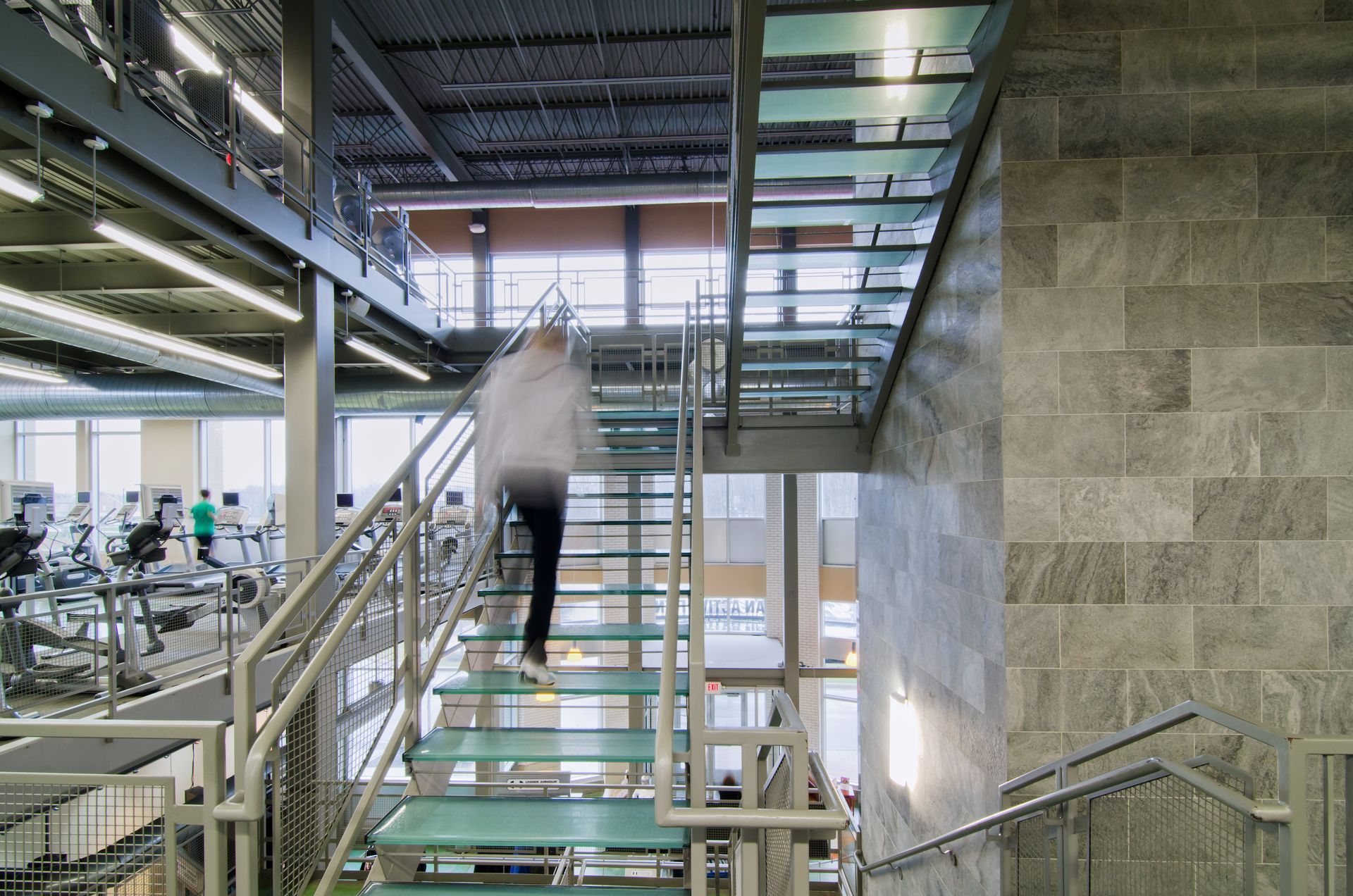 Person blurry climbing stairs in a modern gym, with glass steps, metal railings, and exercise equipment visible.