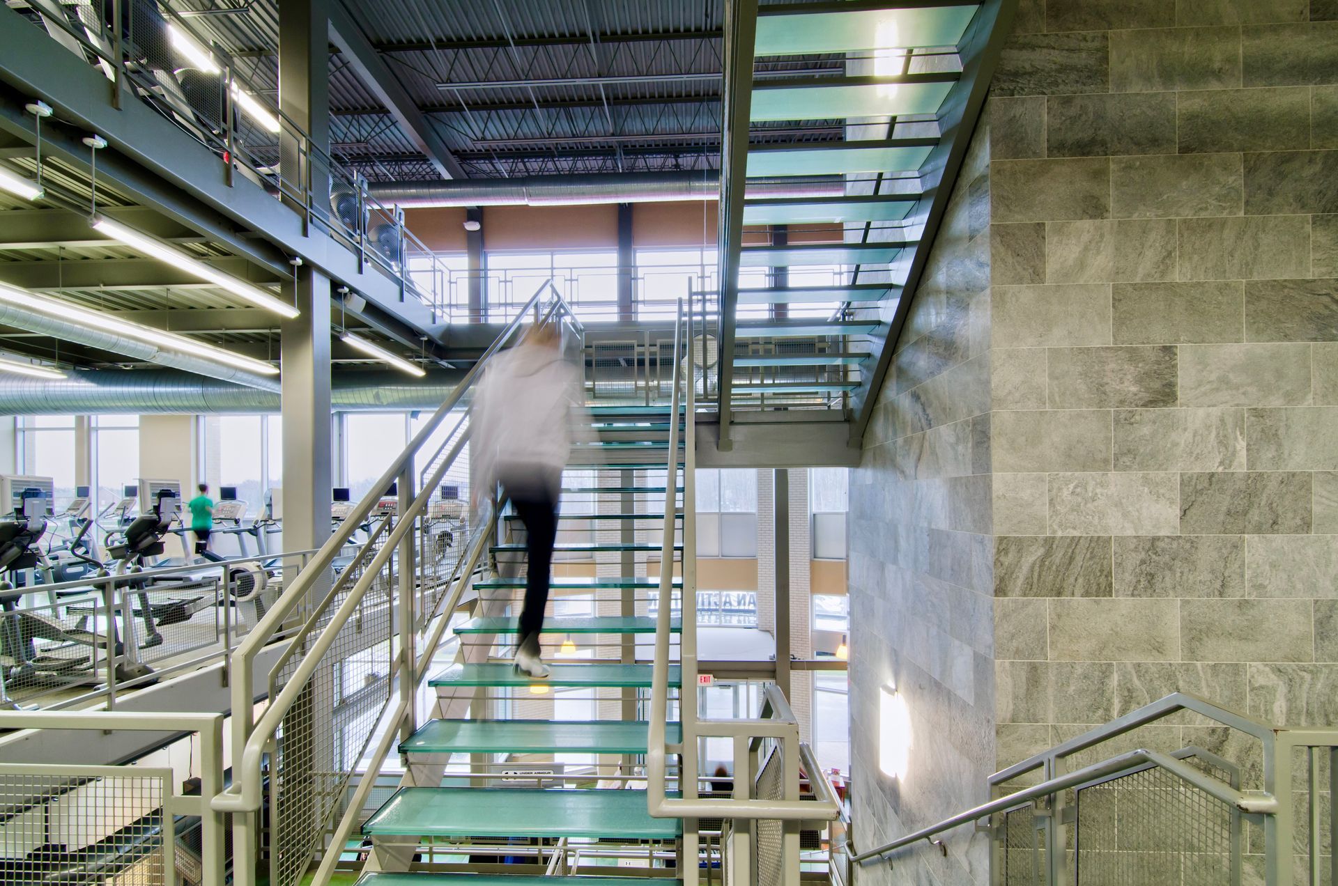 A blurred person ascends modern glass stairs in a gym, with exercise equipment visible on the lower level.