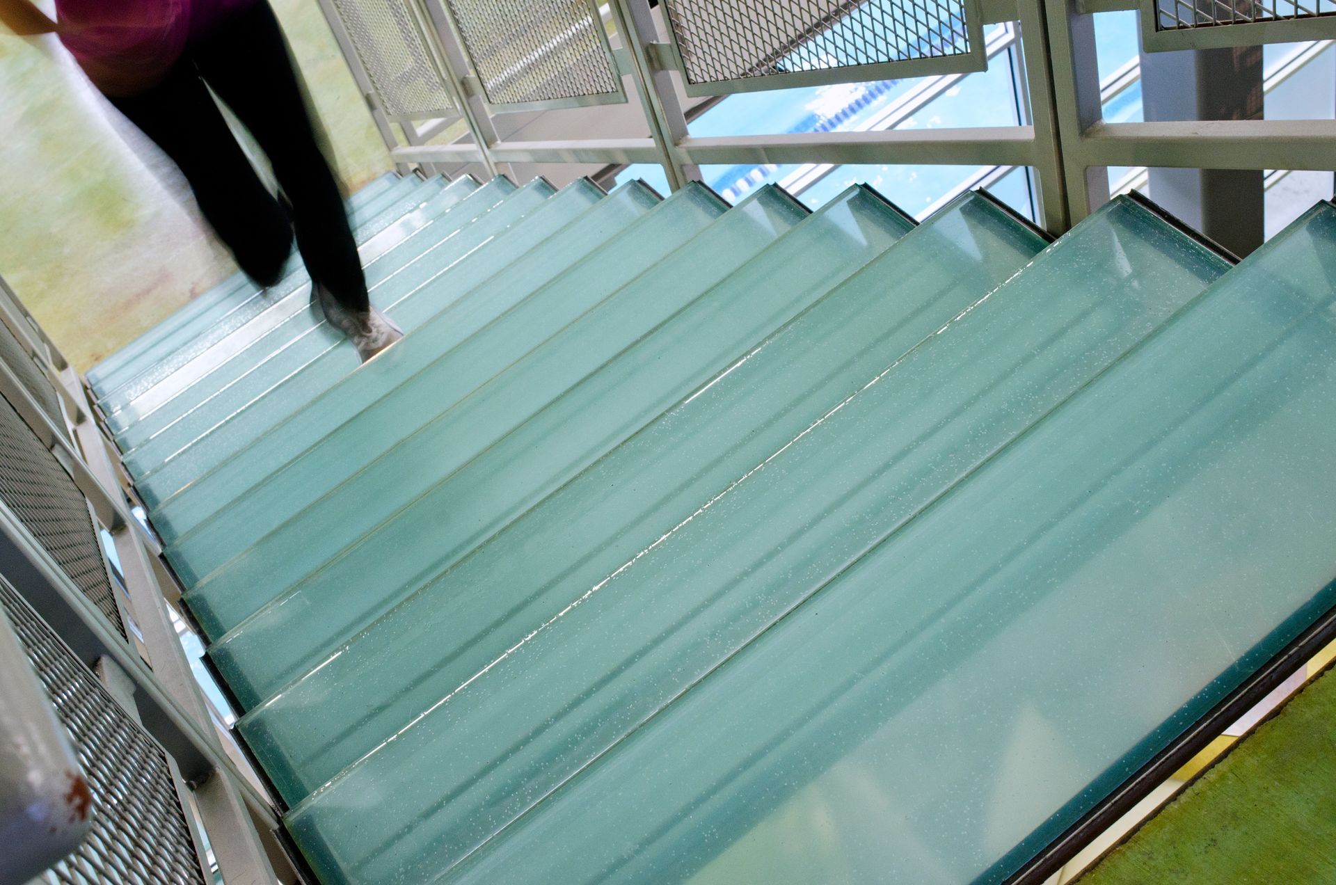 Person walking down glass stairs, legs visible. Metallic railing and blue sky visible.
