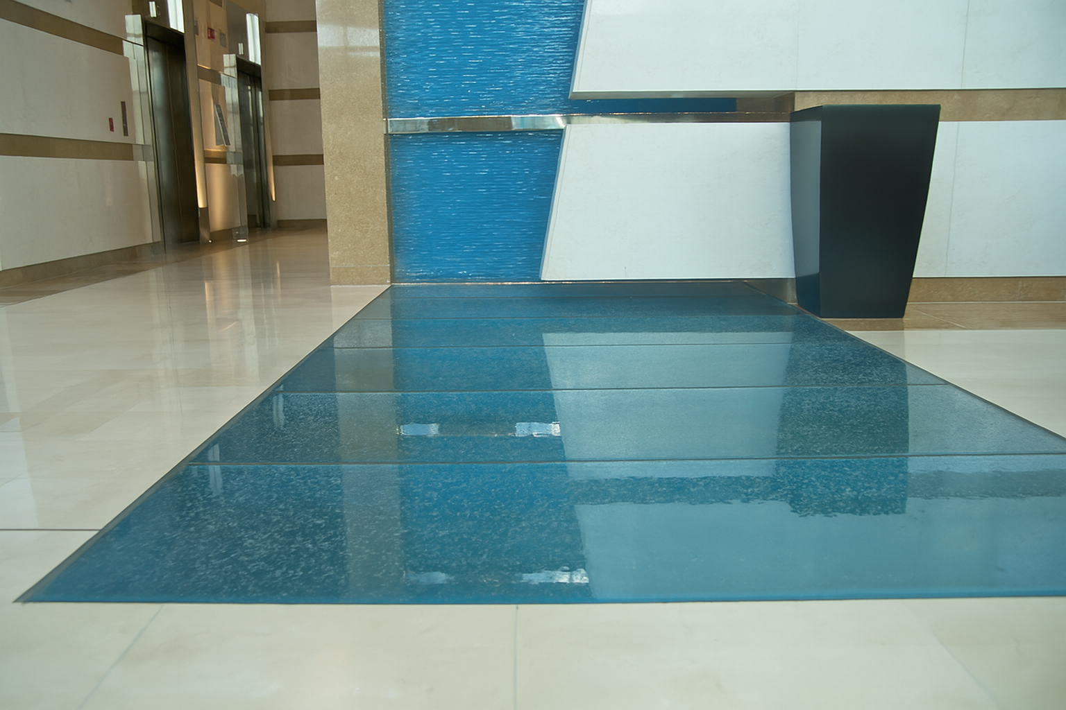 Blue textured glass floor inlay in an office lobby with patterned white walls, beige and black accents, and elevator doors.