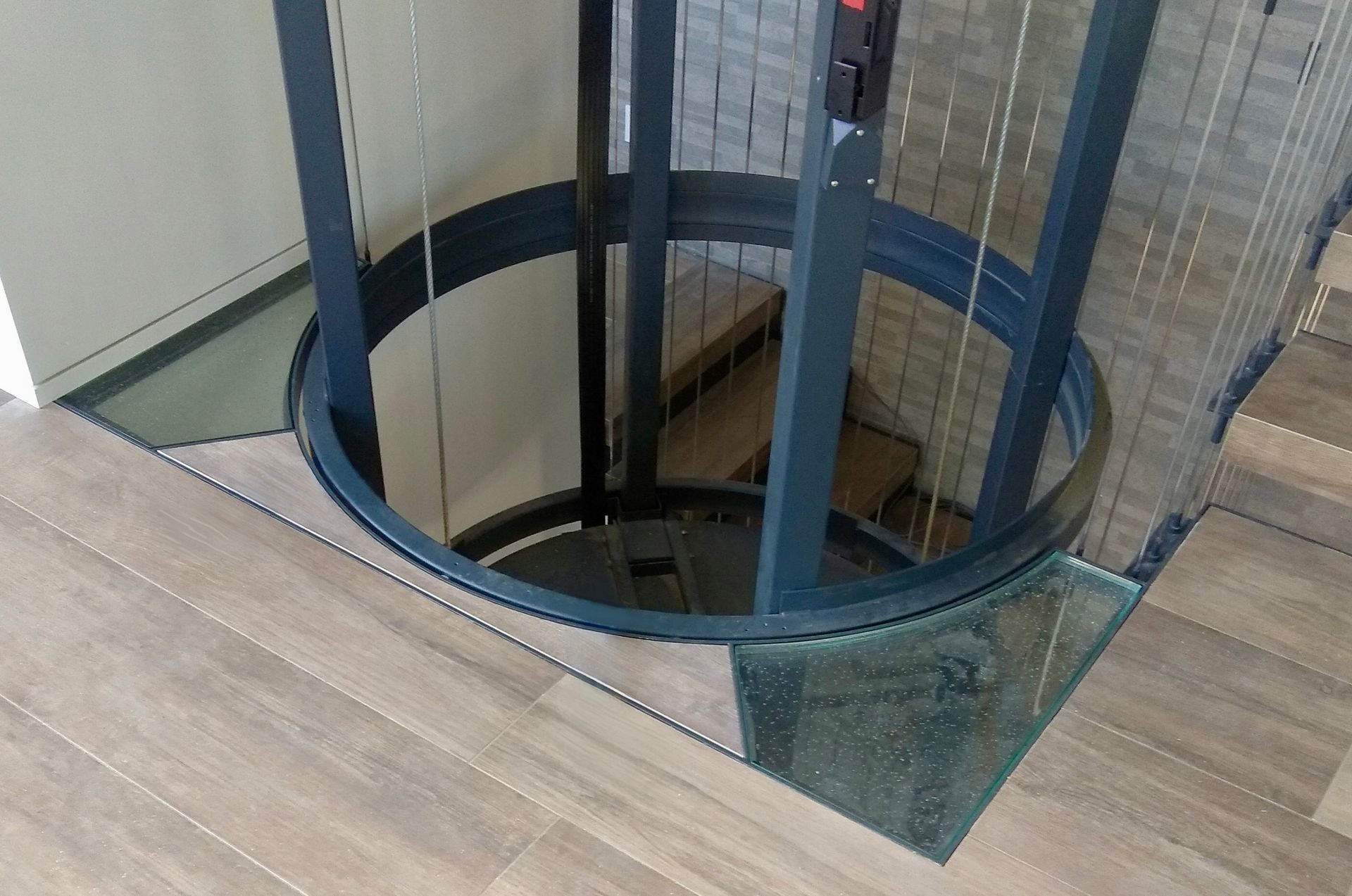 A circular residential elevator with a dark grey frame is installed in a floor with wood-look tile and glass floor panels.