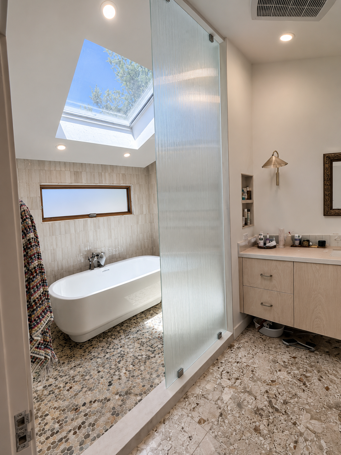 Modern bathroom with freestanding tub, glass shower divider, skylight, and light wood vanity