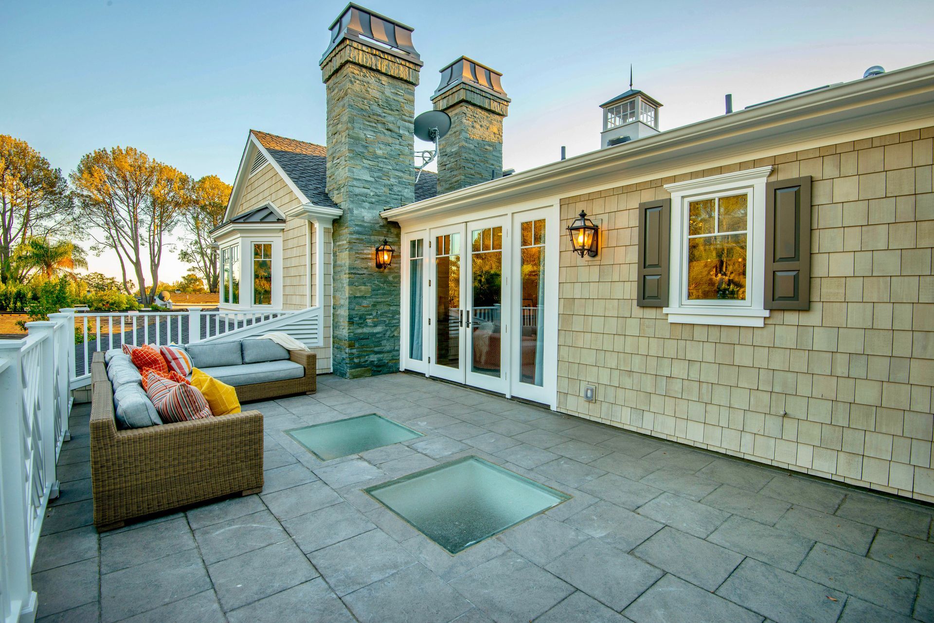 Patio with wicker sofa, glass panels, and French doors in a beige shingled house; trees in the background.