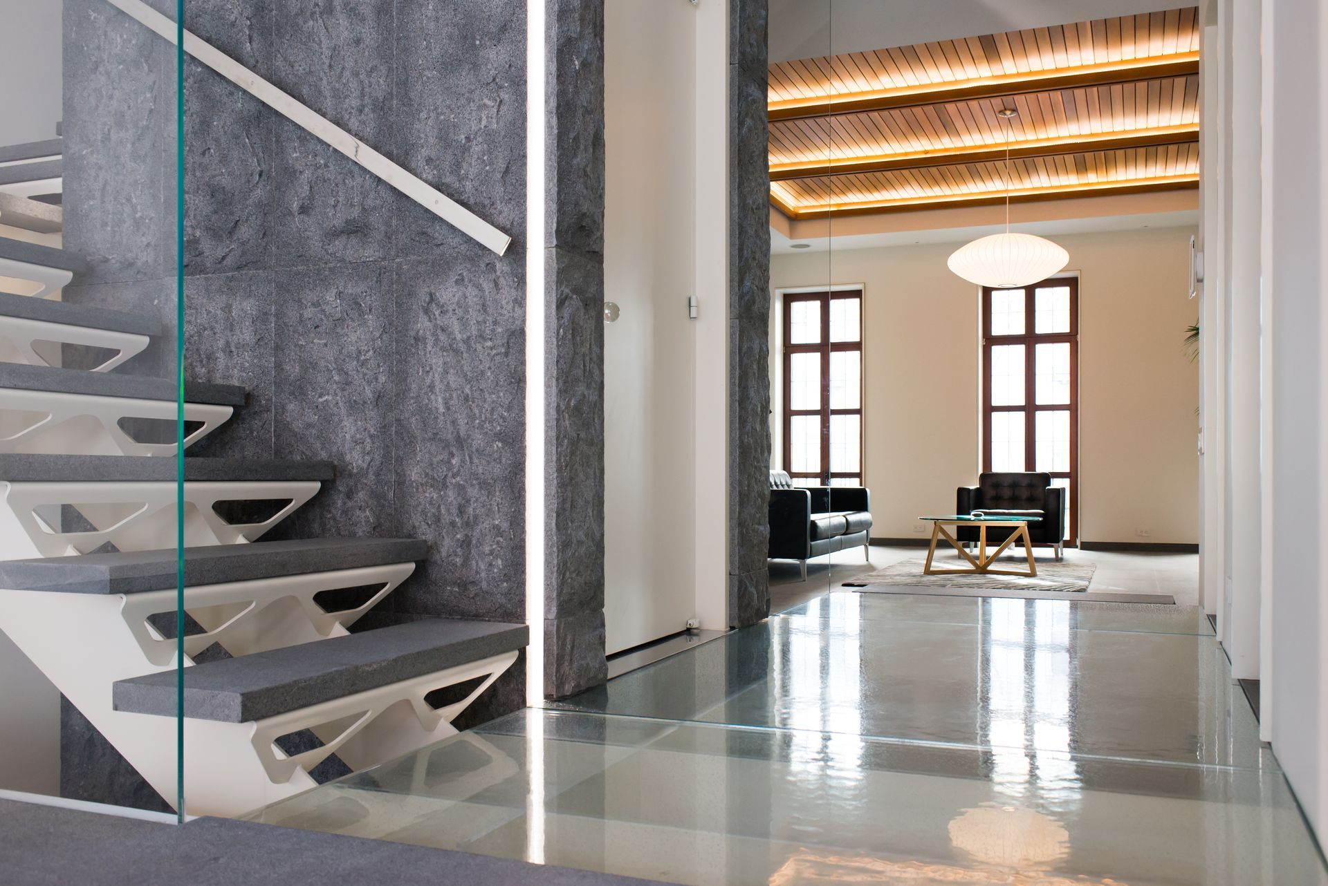 Modern interior with a floating staircase, gray stone wall, and polished floor leading to a bright living area.