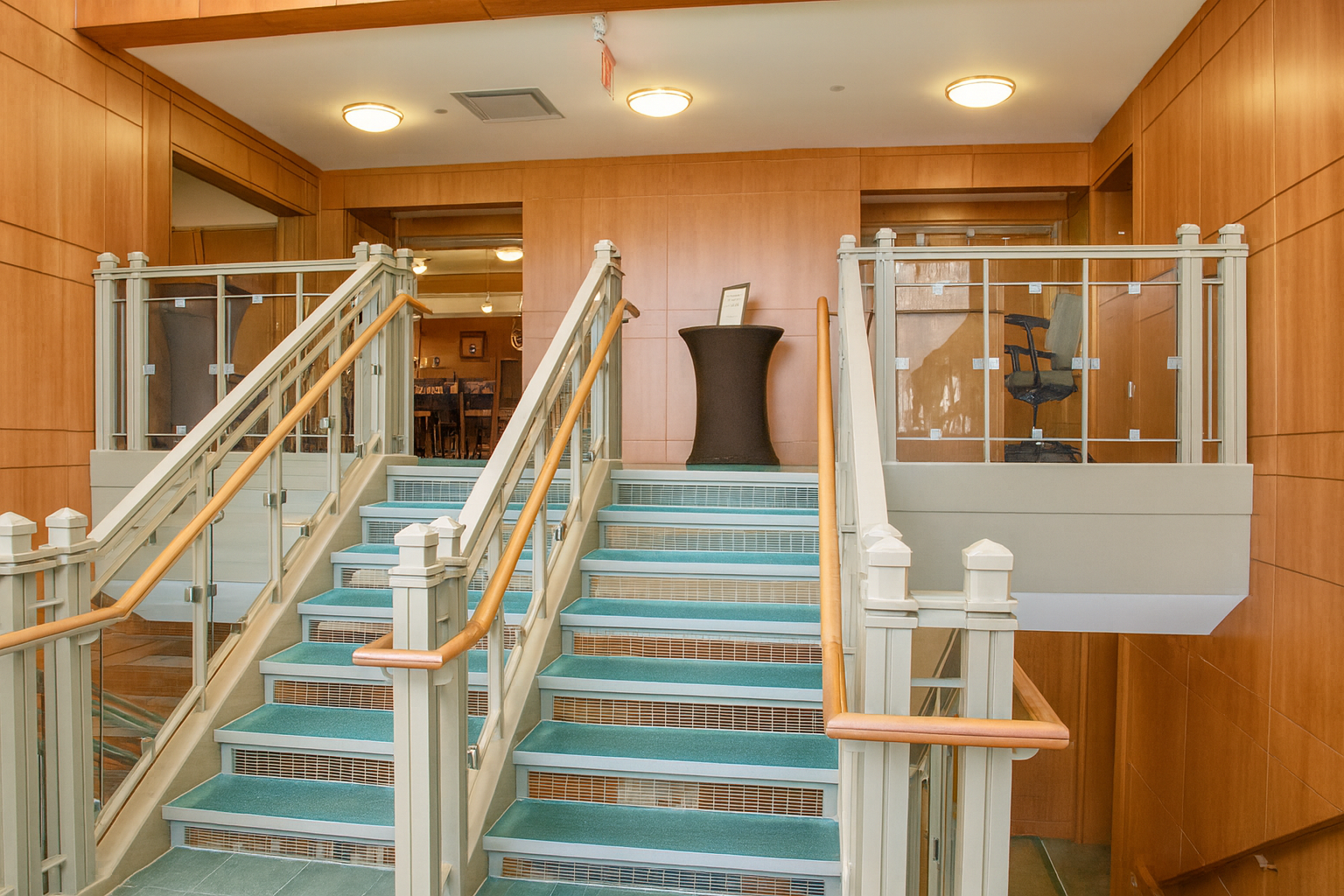 Wooden staircase with turquoise carpet and glass railings, leading to a dining area, small table in between.