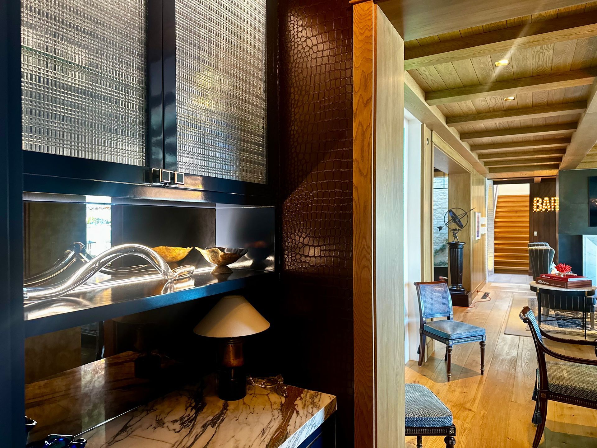 Dark built-in bar with decorative glass doors, leading to a hallway with hardwood floors and stairs.
