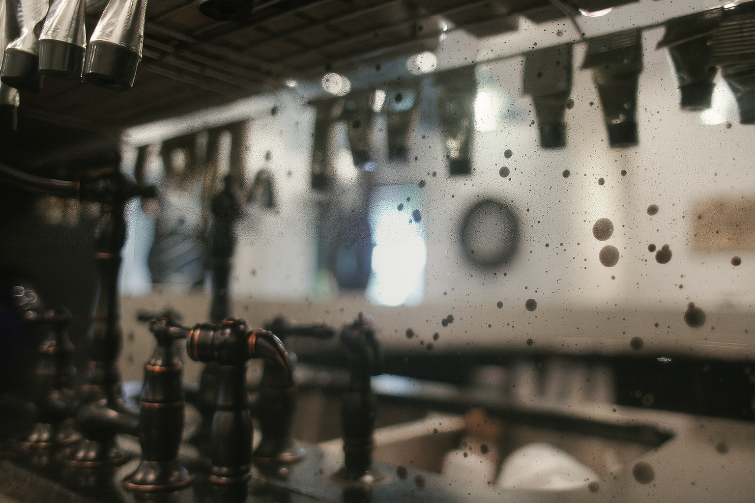 A weathered, speckled mirror reflects a faucet and blurry interior, suggesting a vintage washroom.