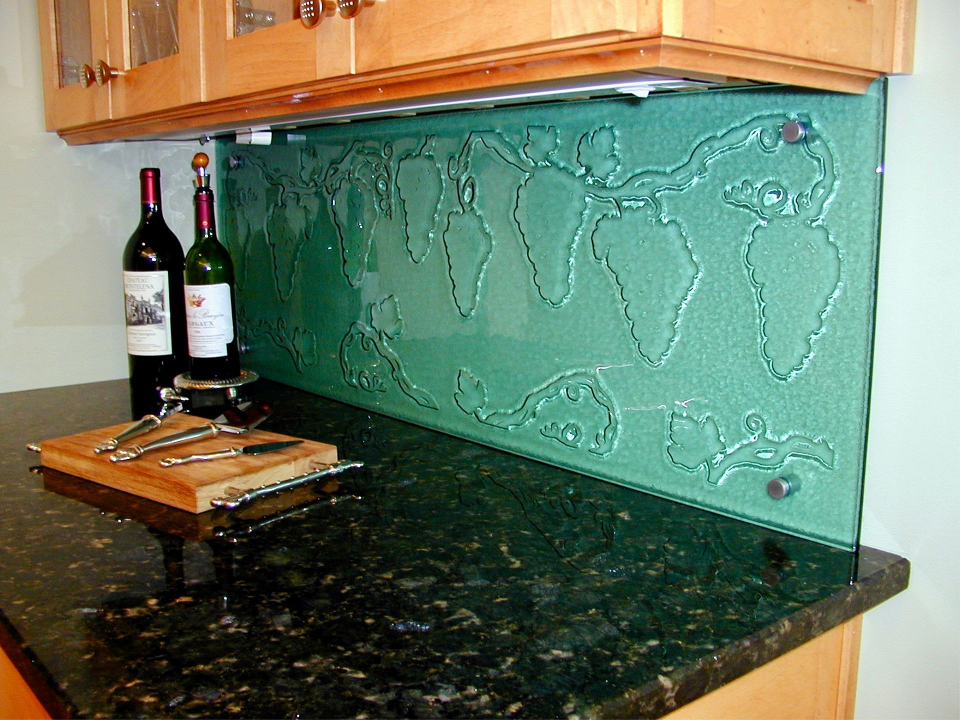 A green glass kitchen backsplash featuring an etched grape vine design, mounted above a dark granite countertop with wine.