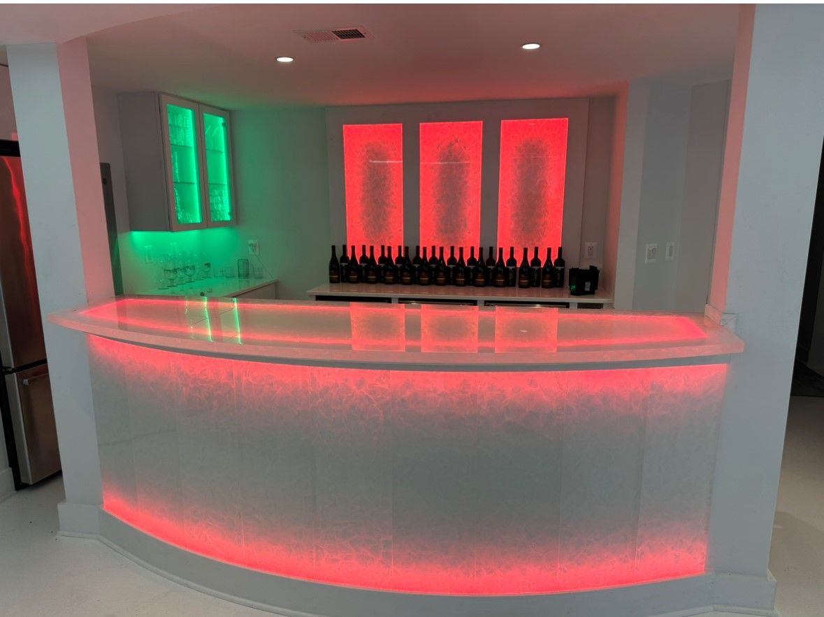 A curved home bar featuring a white countertop with red LED under-lighting and glowing red and green wall panels.