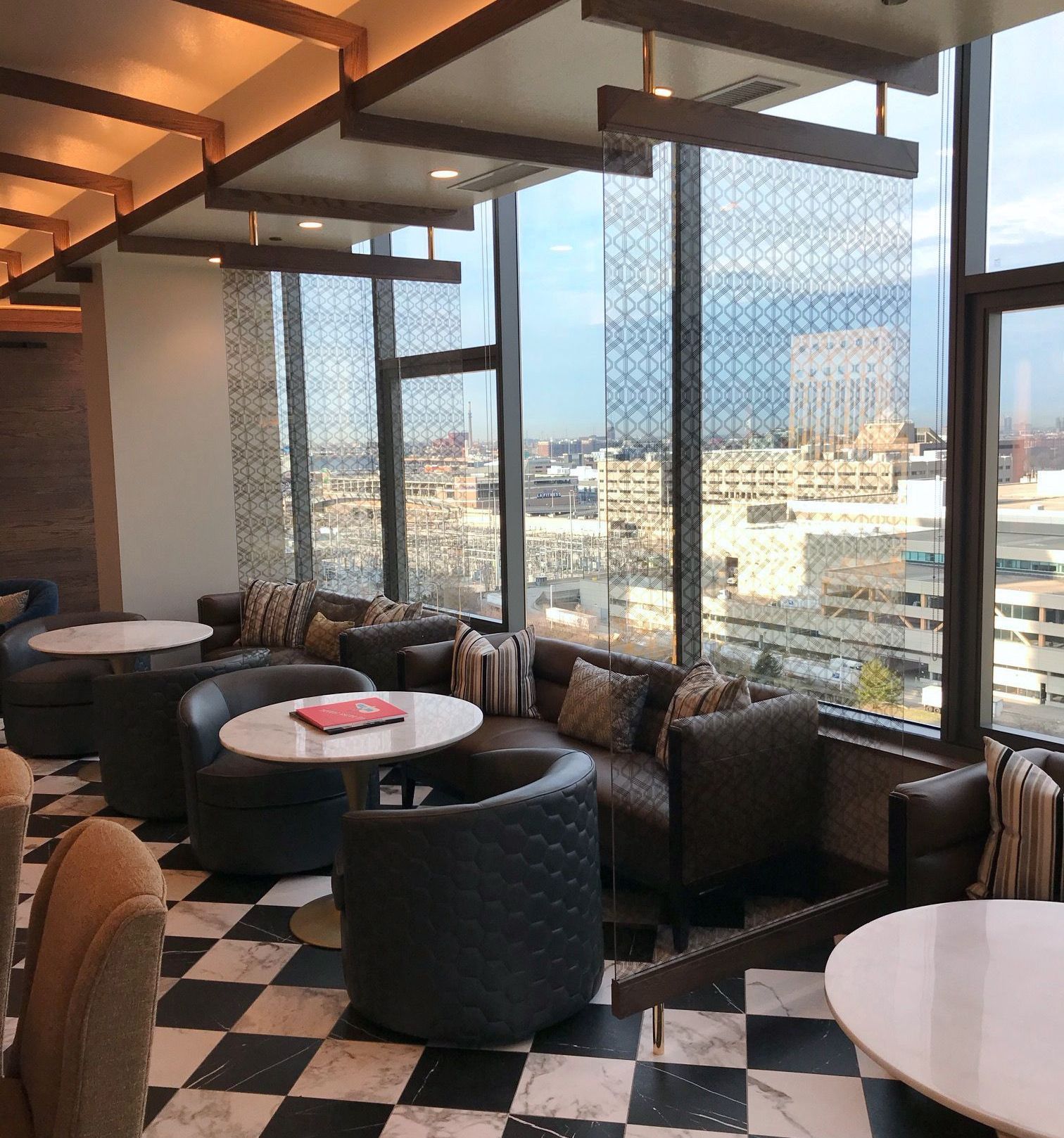Lounge area with large windows overlooking a city. Gray and black checkerboard floor, circular seating.