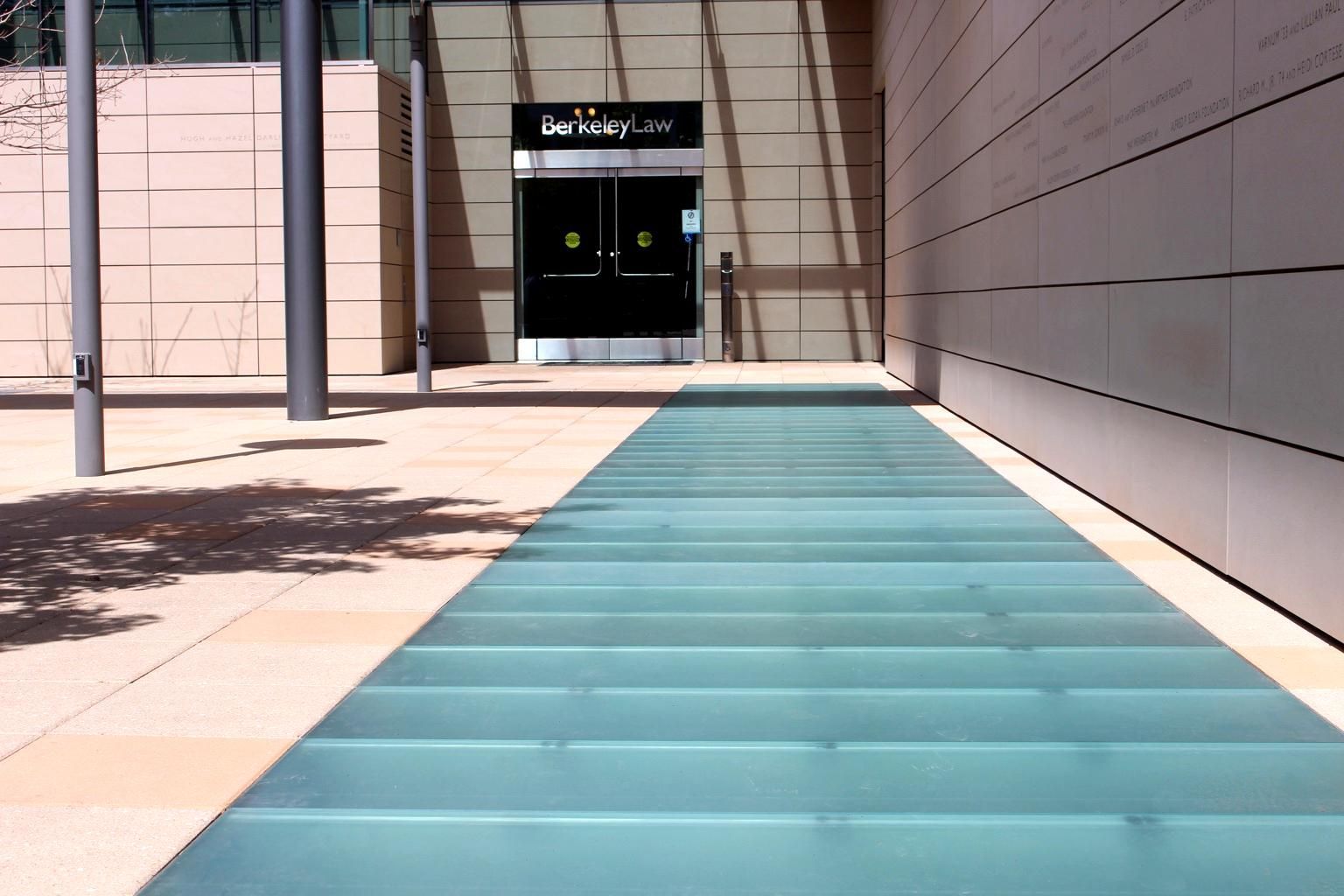 Glass walkway leads to a doorway, between tan buildings and posts; sunny outdoor scene.