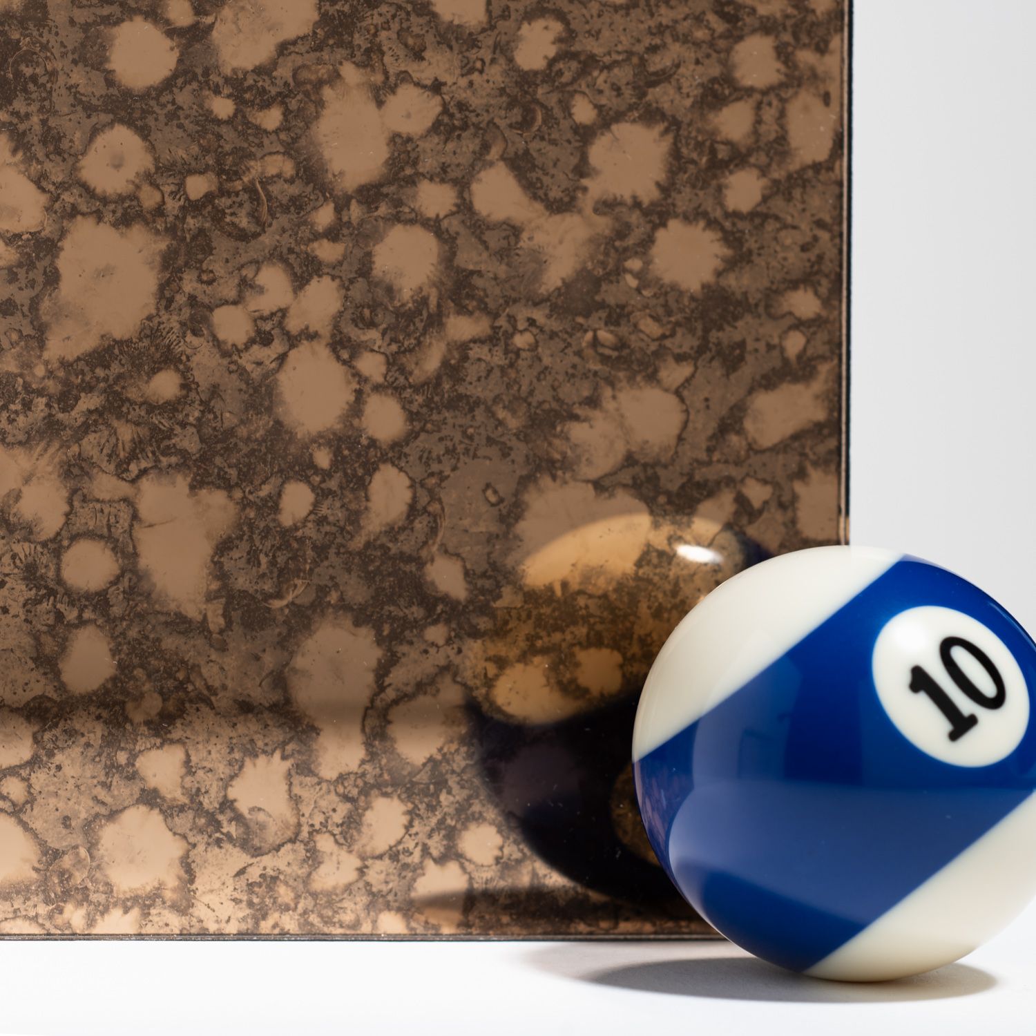 A blue and white number 10 billiard ball rests in front of a rectangular piece of bronze-colored antique mirror glass.