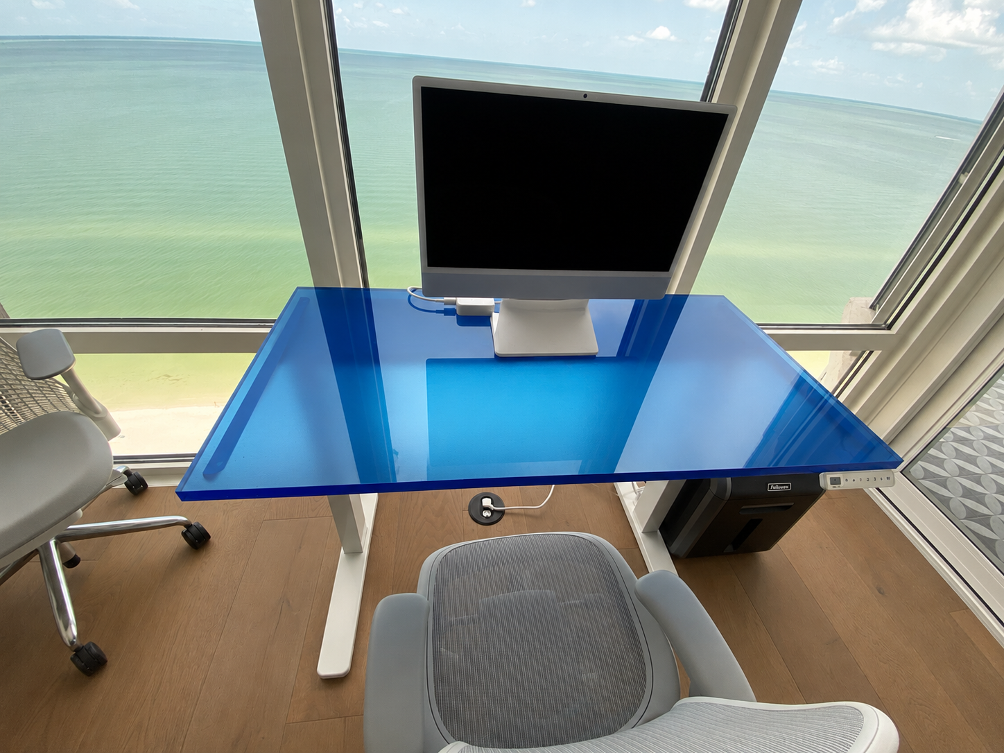 Blue glass desk with a monitor by a window, viewed from above.