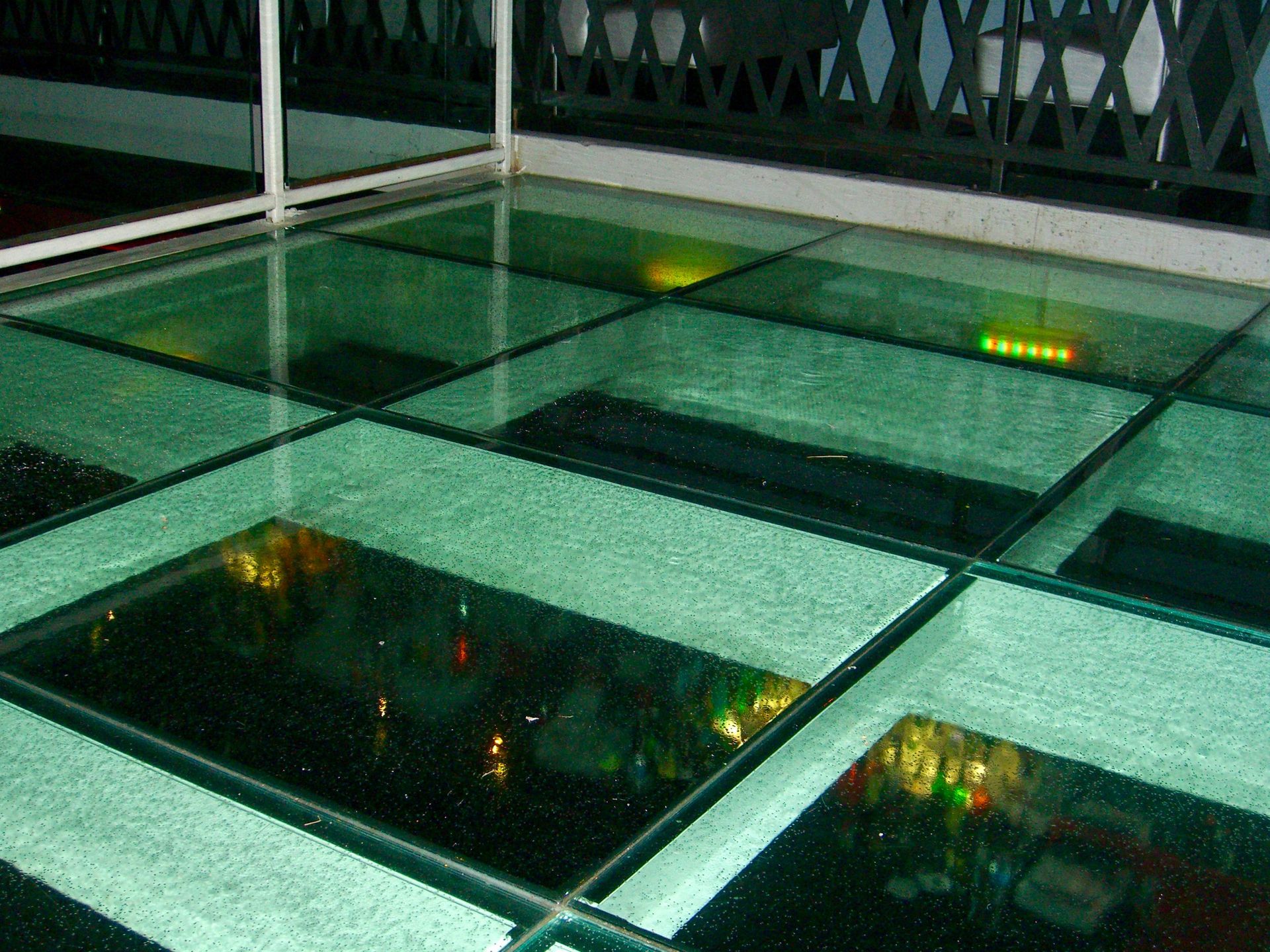Glass floor panels on a balcony overlooking a city at night. Green-tinted glass reflects lights below.