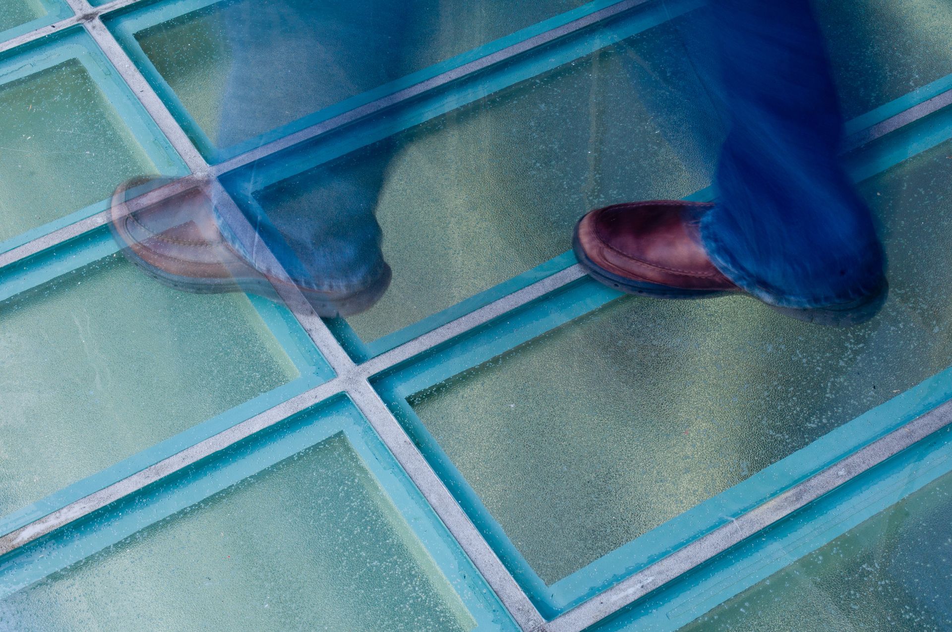 Person's feet in brown shoes walking on a clear glass walkway with turquoise borders.