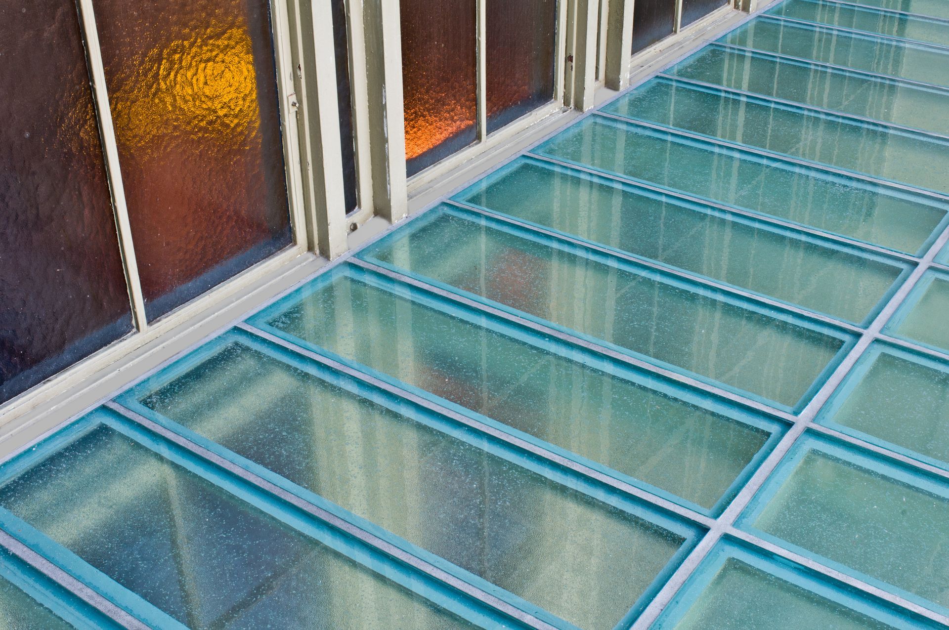 A glass floor panel system next to a wall with large windows, showing a repeating grid structure.
