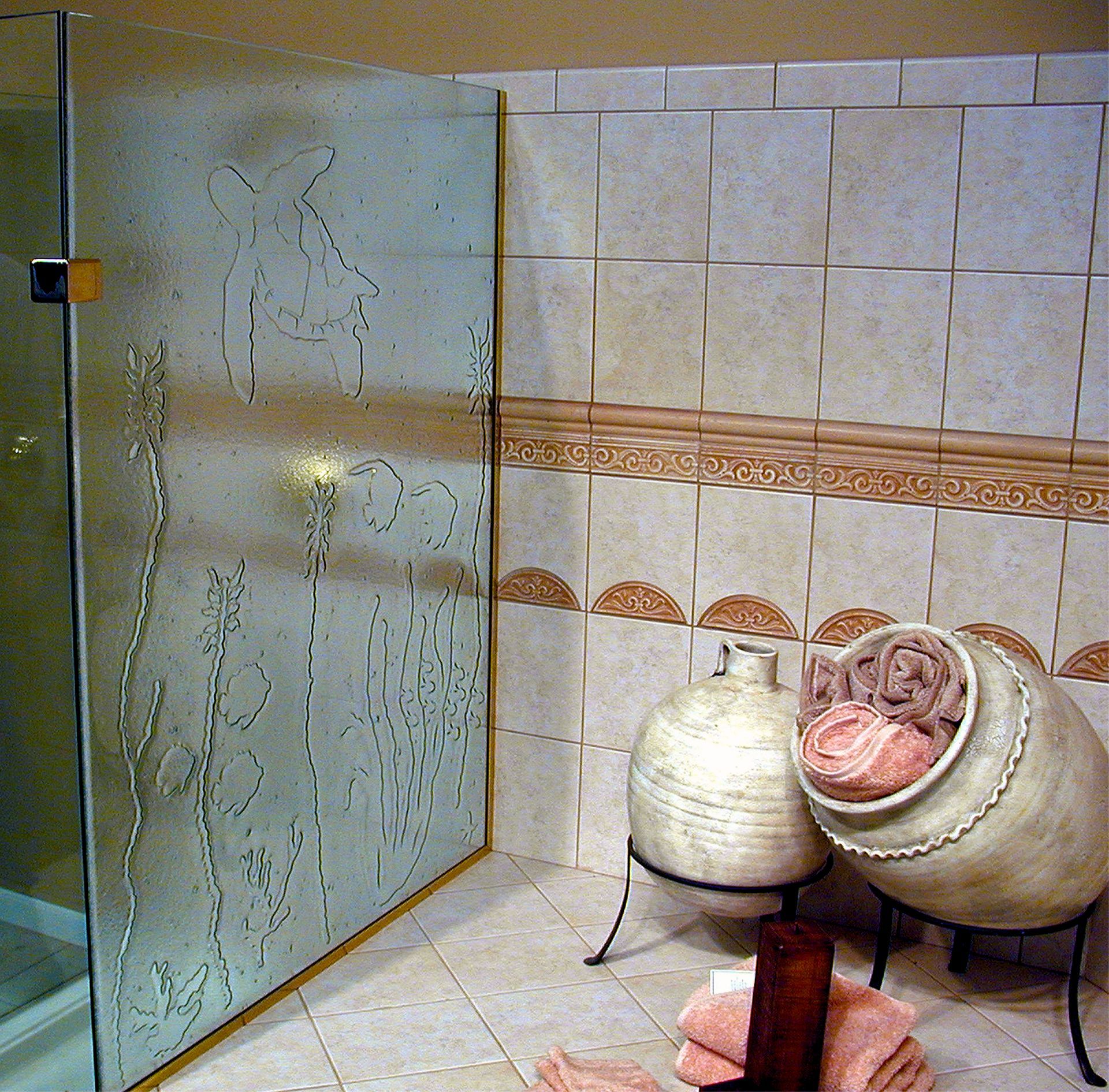 A glass shower door etched with fish and plants beside tiled walls, two decorative pottery vessels, and pink towels.