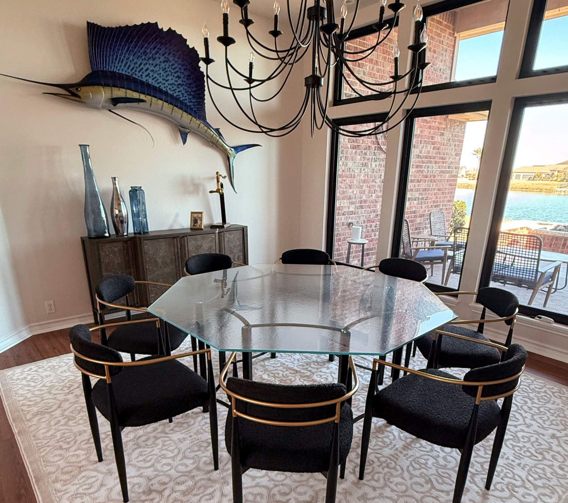 Dining room with glass-top table, black chairs, chandelier, and a large blue fish sculpture on the wall.