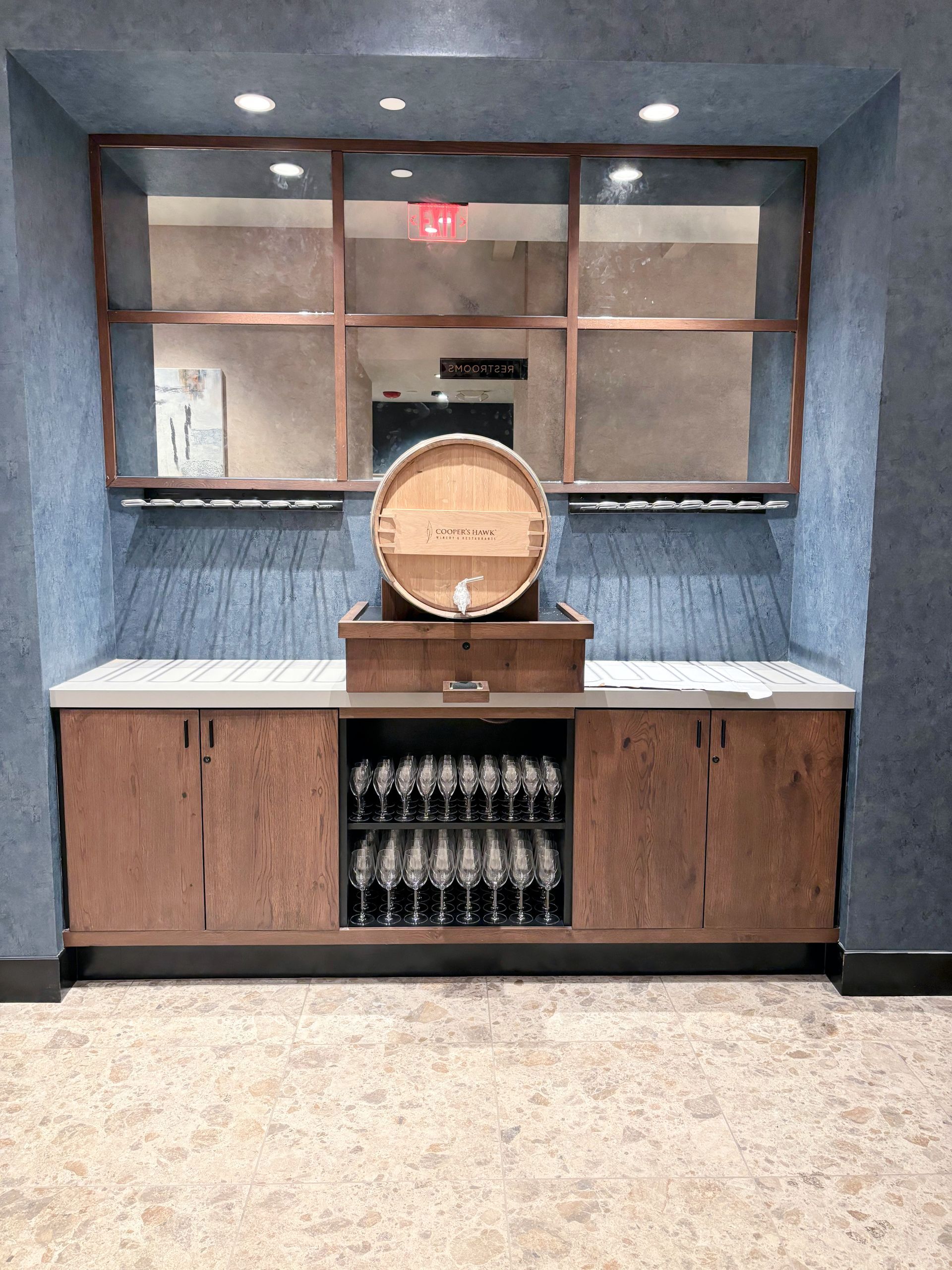 Wooden wine barrel on cabinet in a blue-walled alcove with glassware and a mirrored backdrop.