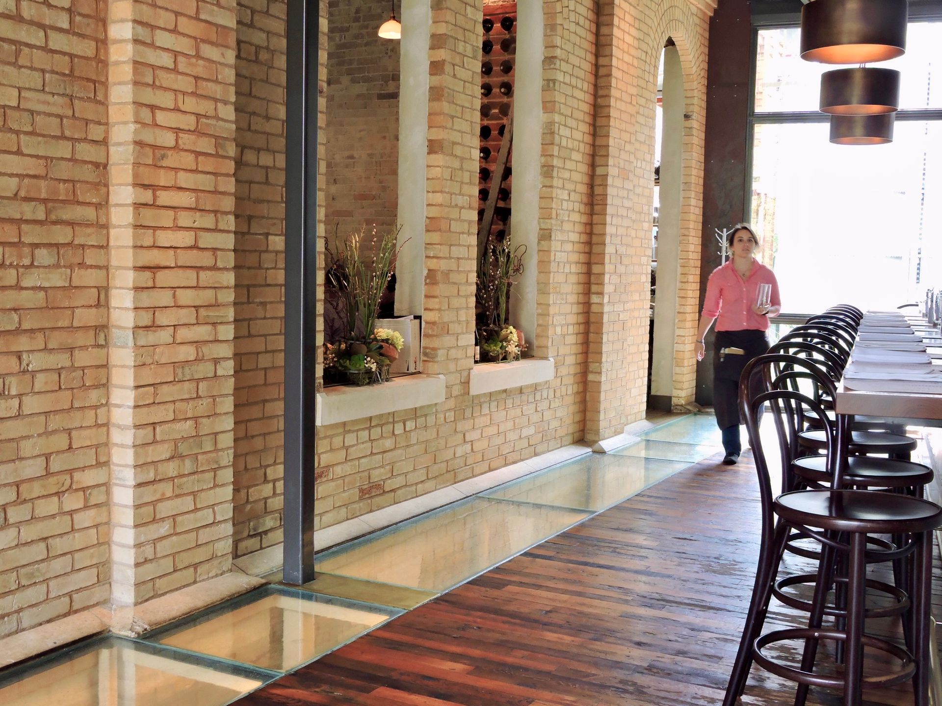 Restaurant interior with brick walls, glass floor, wooden bar, server walking.