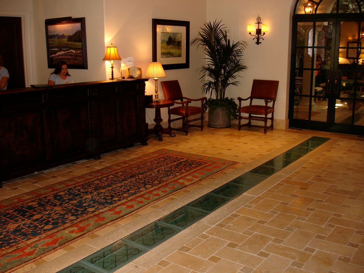 Reception area with a wooden desk, two chairs, a potted plant, and a decorative rug on a tiled floor.