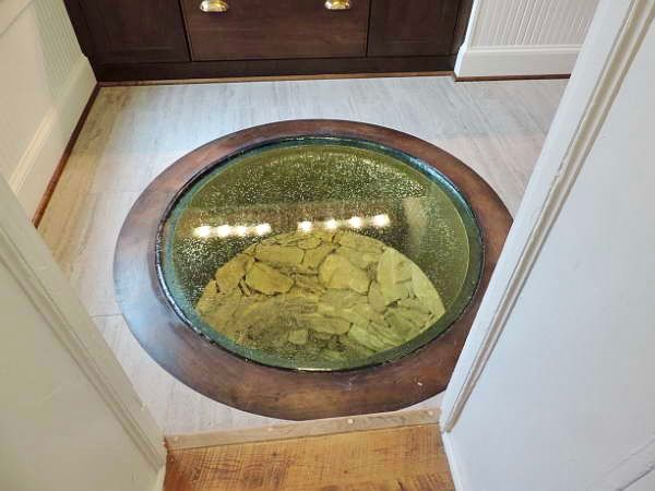 A circular glass floor insert embedded in light-colored flooring, revealing a shallow, rock-filled space below.