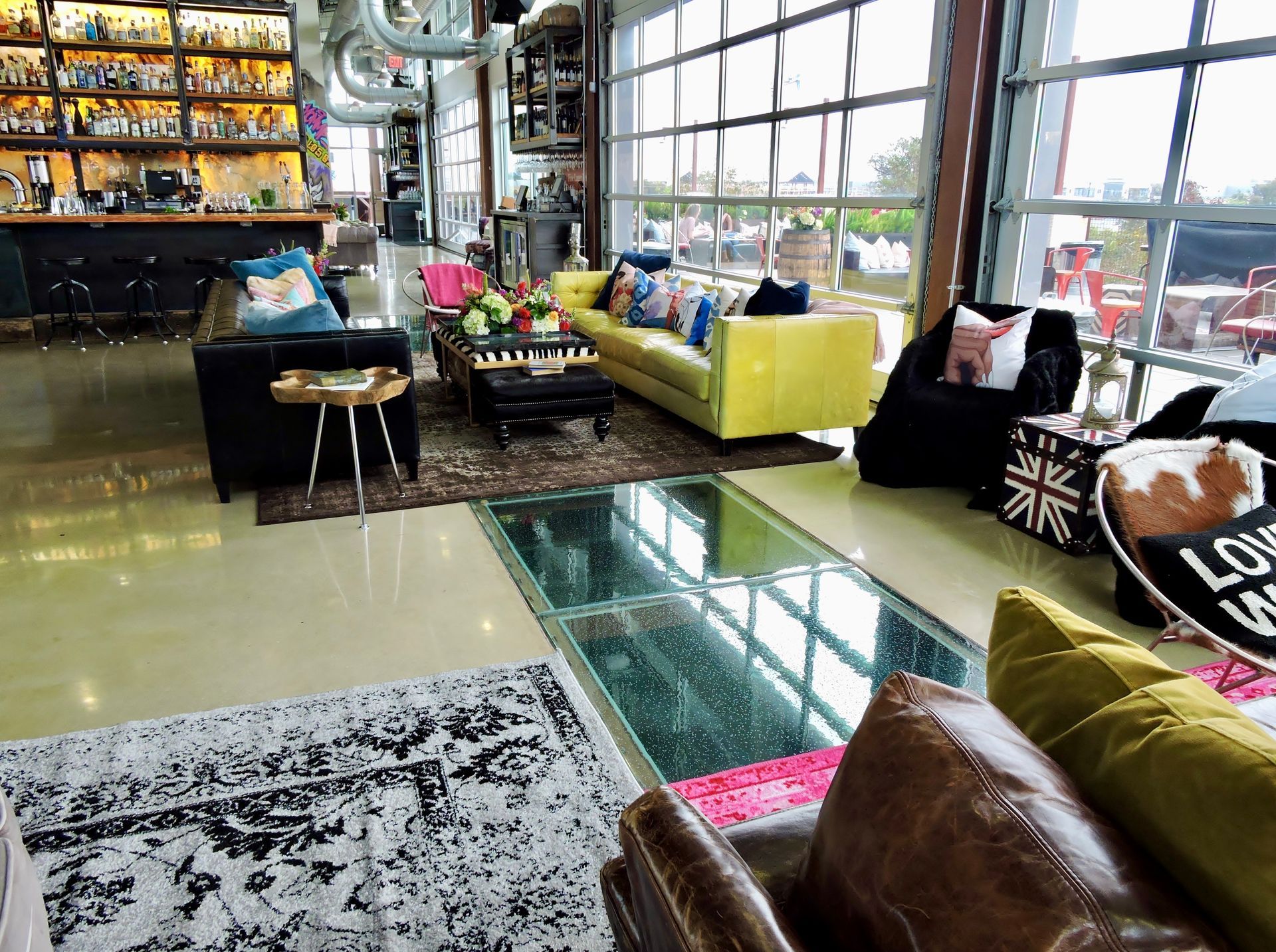 Lounge area with seating: black, yellow, and brown sofas. Large windows offer an outdoor view. Bar in the background.