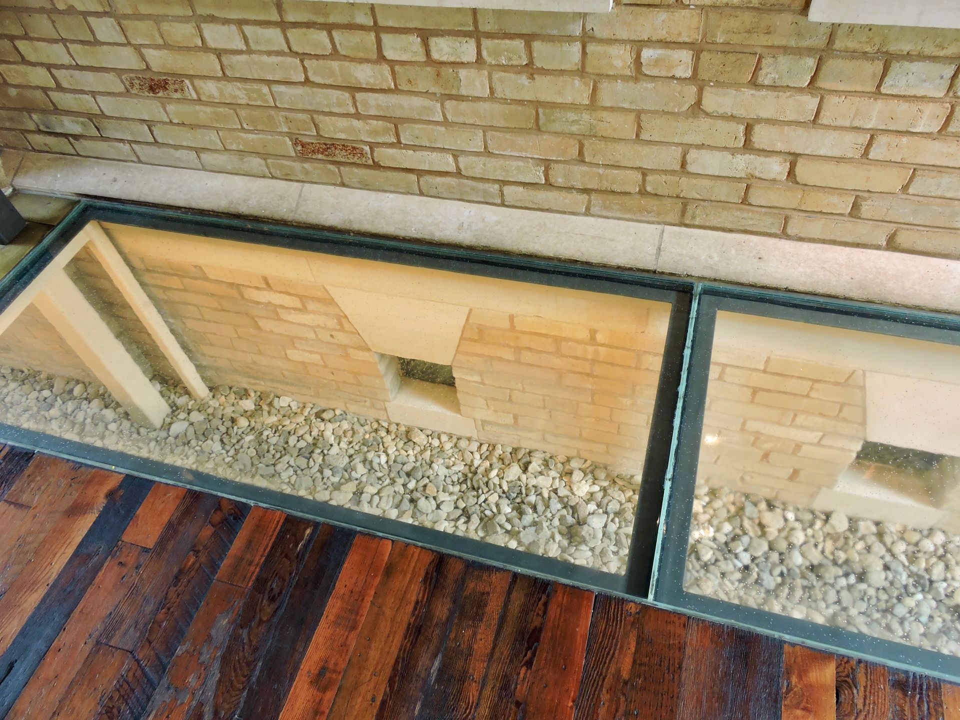 Glass floor panels reveal brick foundation and gravel, set against wood flooring and a brick wall.