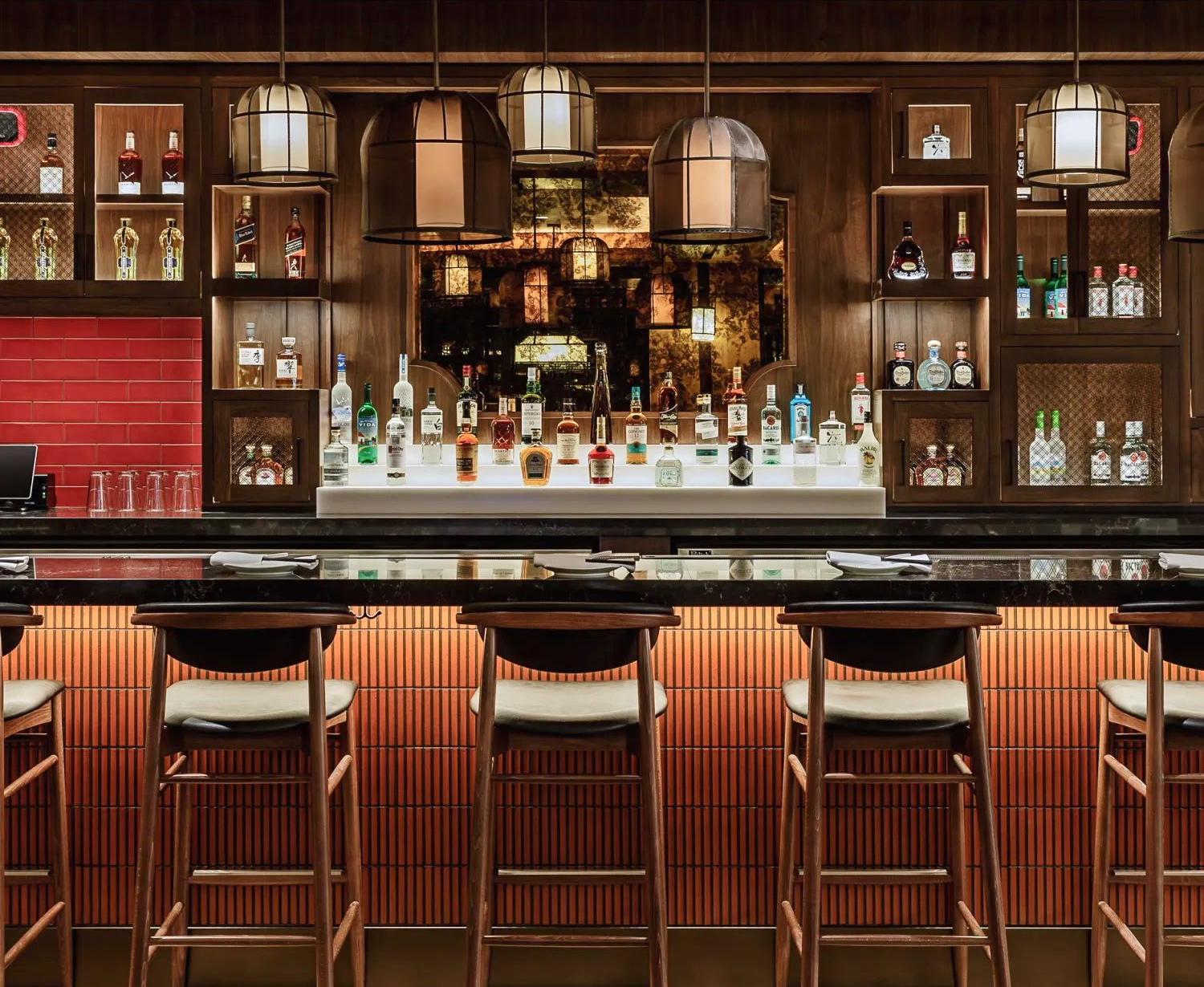 Cozy bar with backlit wood paneling, shelves of liquor, and stools. Warm lighting, dark wood, and bottles on display.