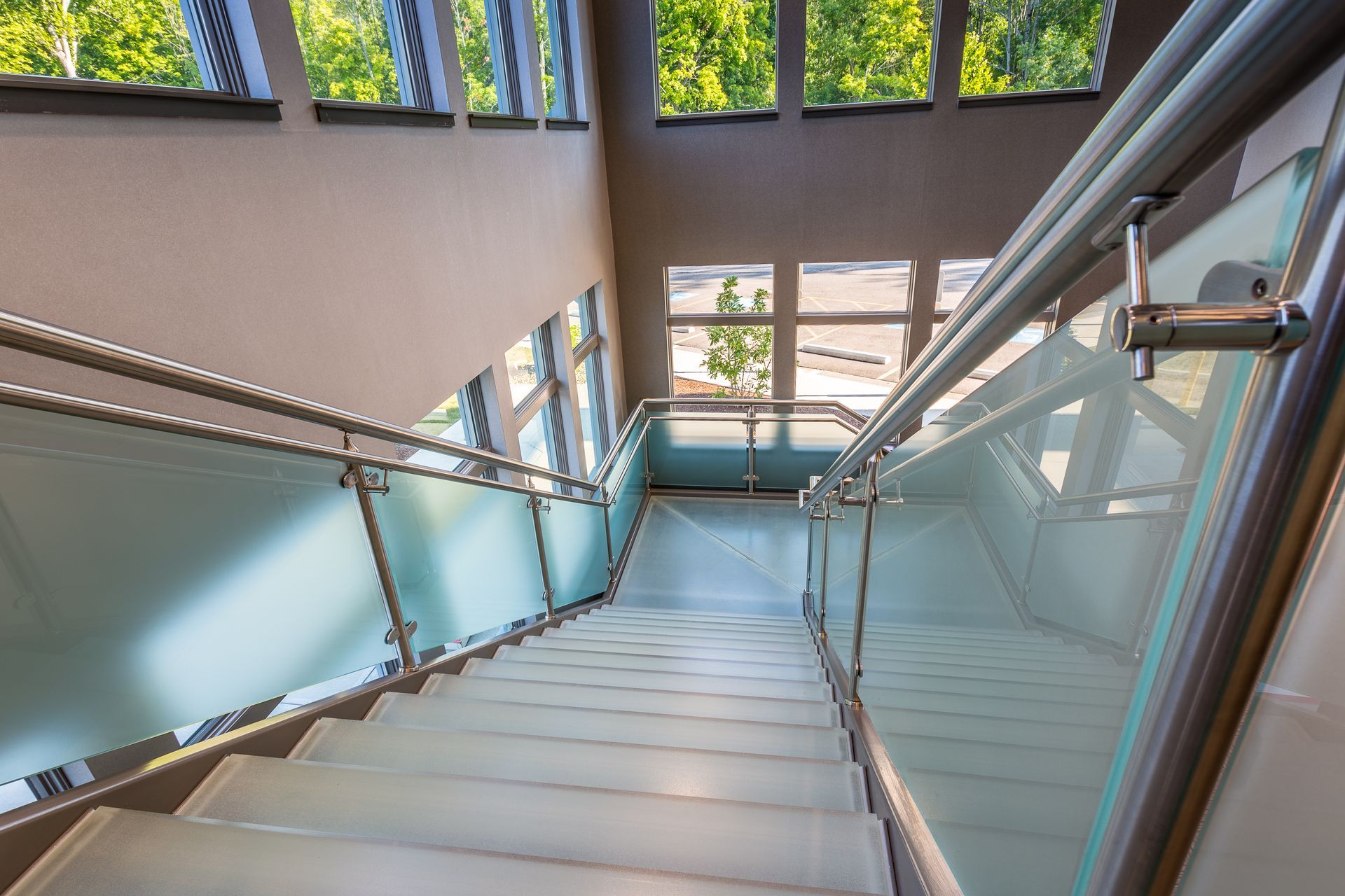 A high-angle view looking down a modern staircase with glass steps and glass railings featuring metallic handrails.