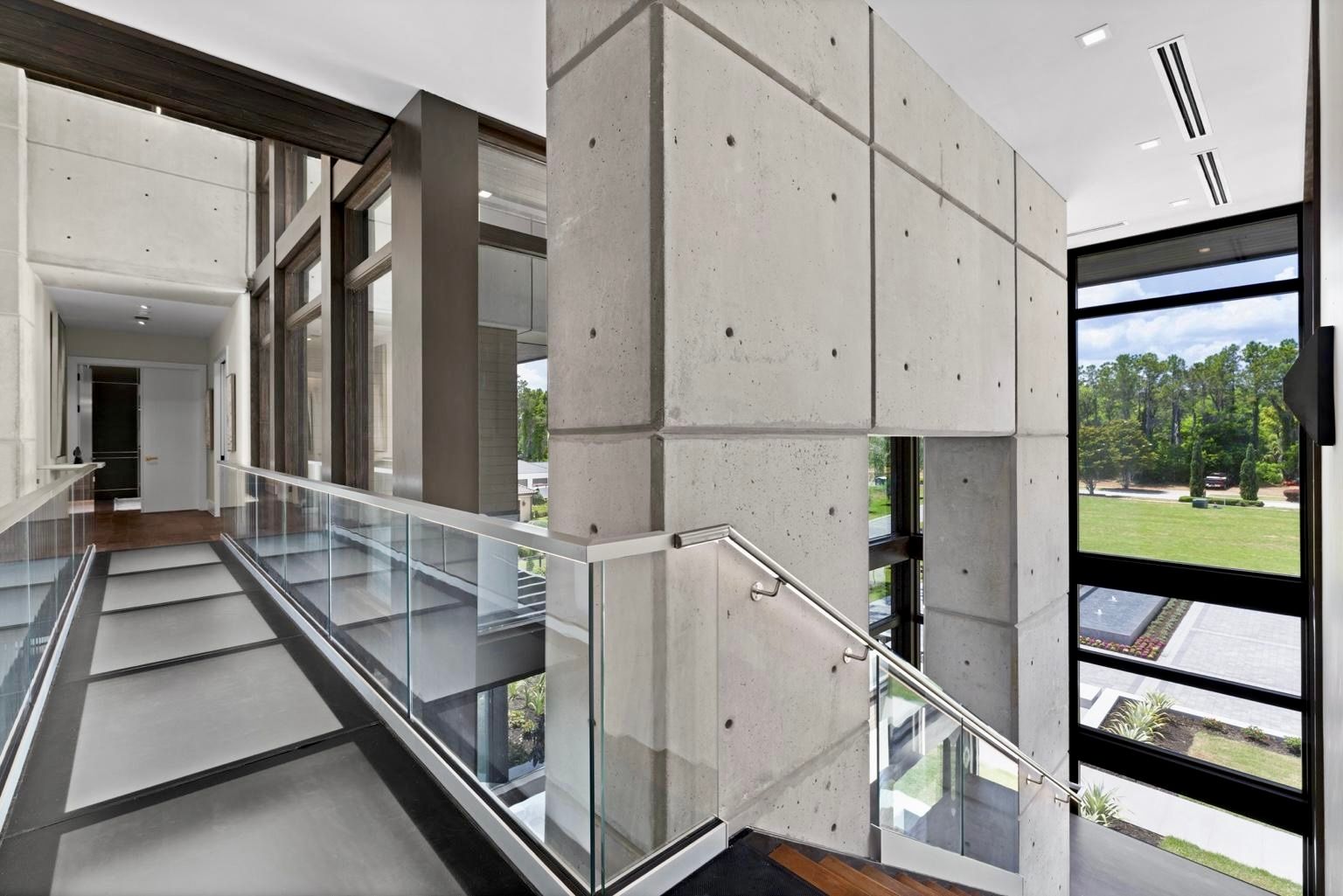 Modern hallway with a concrete wall, glass railings, and floor-to-ceiling windows looking out onto a grassy yard.