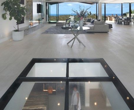 Modern home interior with glass floor panel revealing a staircase below. Ocean view through open doors.