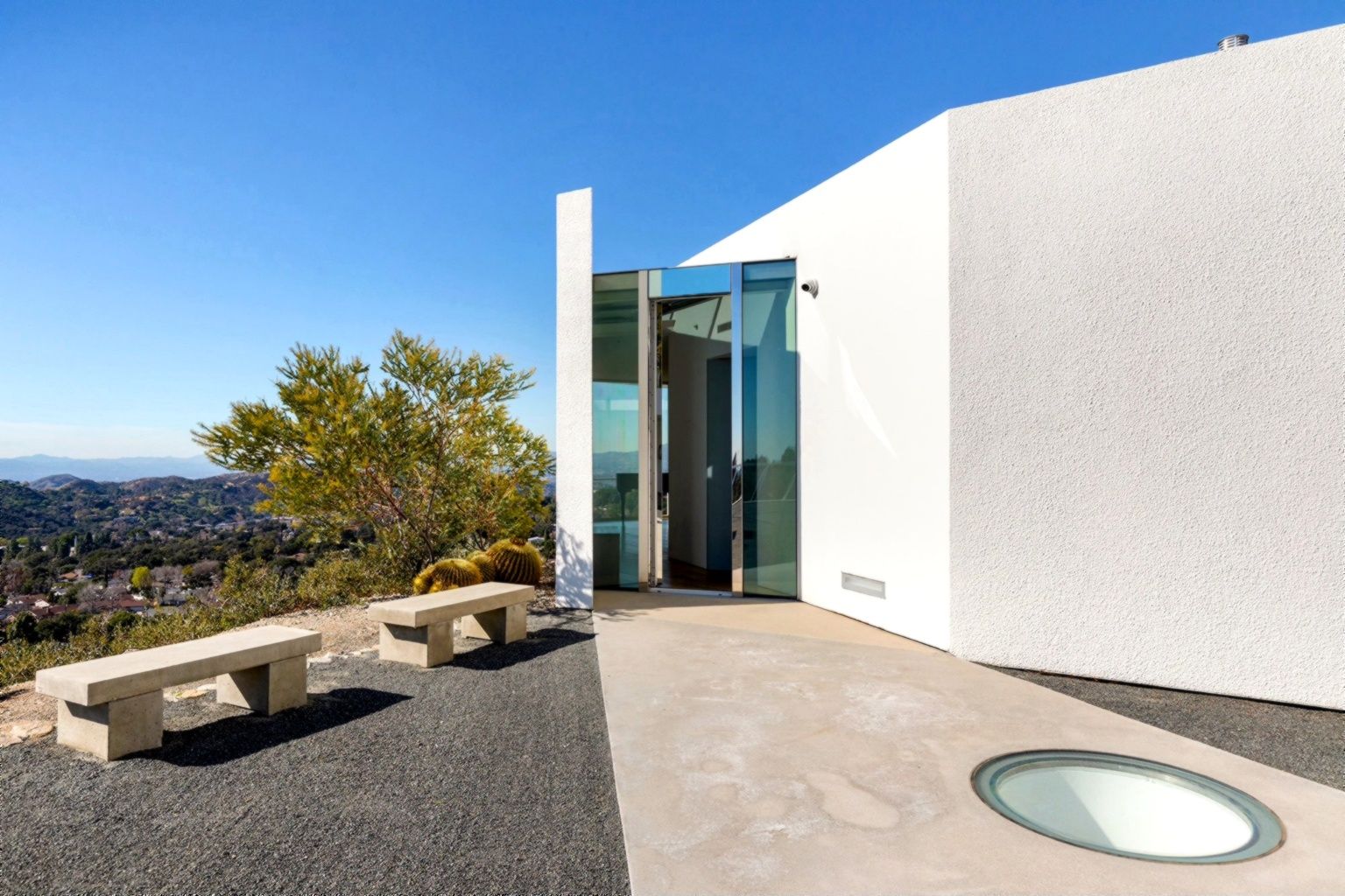 Modern white building with glass doors, stone benches, and a circular glass floor skylight. Overlooks a hillside view.