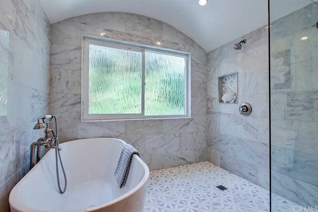 Bathroom with oval tub, marble walls and floor, shower with glass door and window with view.