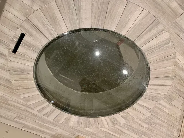 A large, circular, dark glass skylight set into a tiled floor with radiating, light gray wooden-look planks.