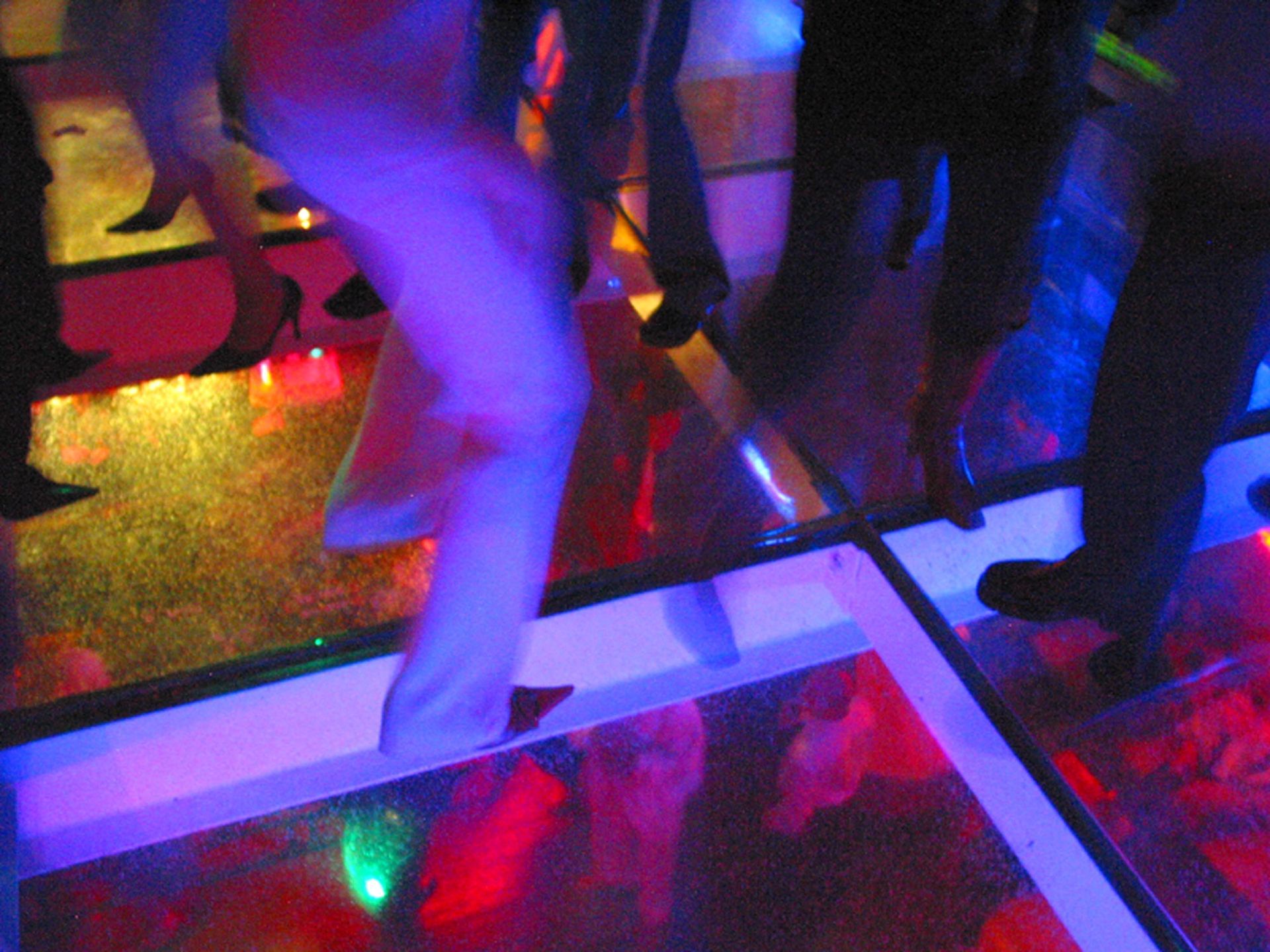 People dancing on a brightly lit, multicolored grid glass dance floor.