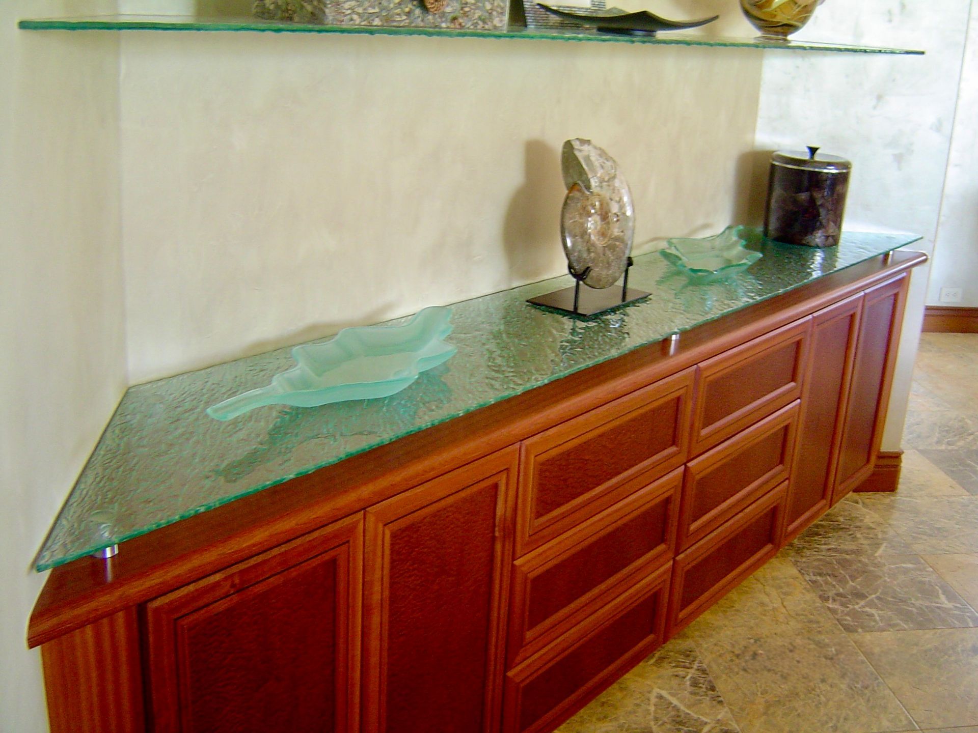 A wooden sideboard with a textured glass top, featuring decorative bowls and an ammonite fossil, set against a wall.