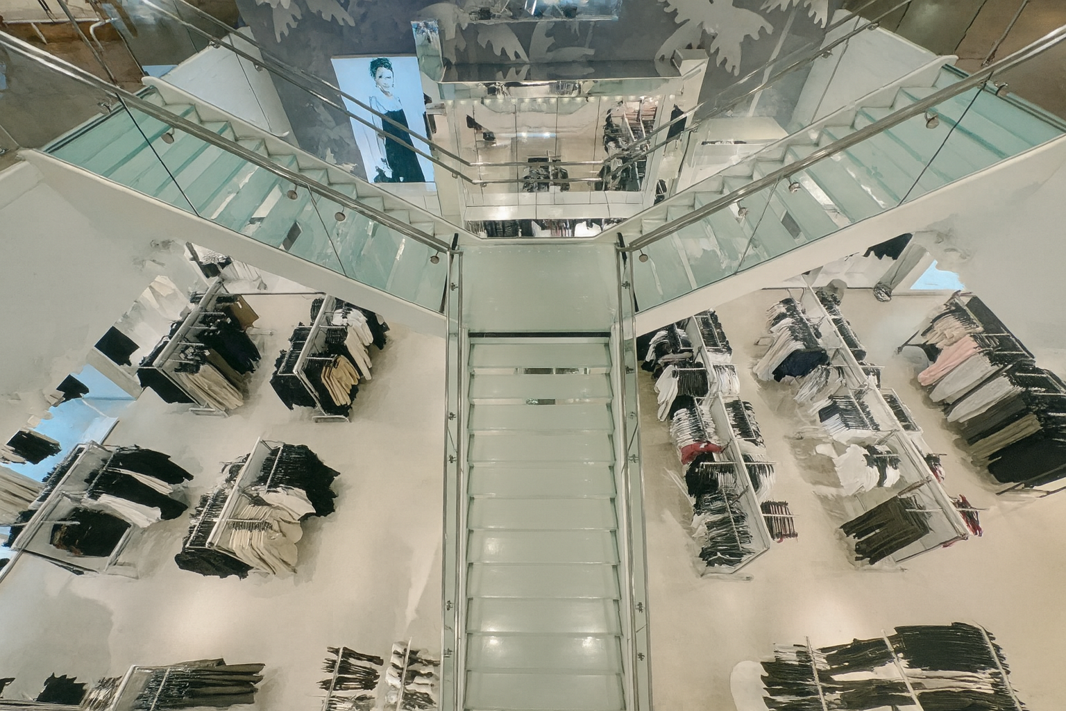 Overhead view of a bright, multi-level clothing store with clothing racks and shoppers; glass railings and staircase.