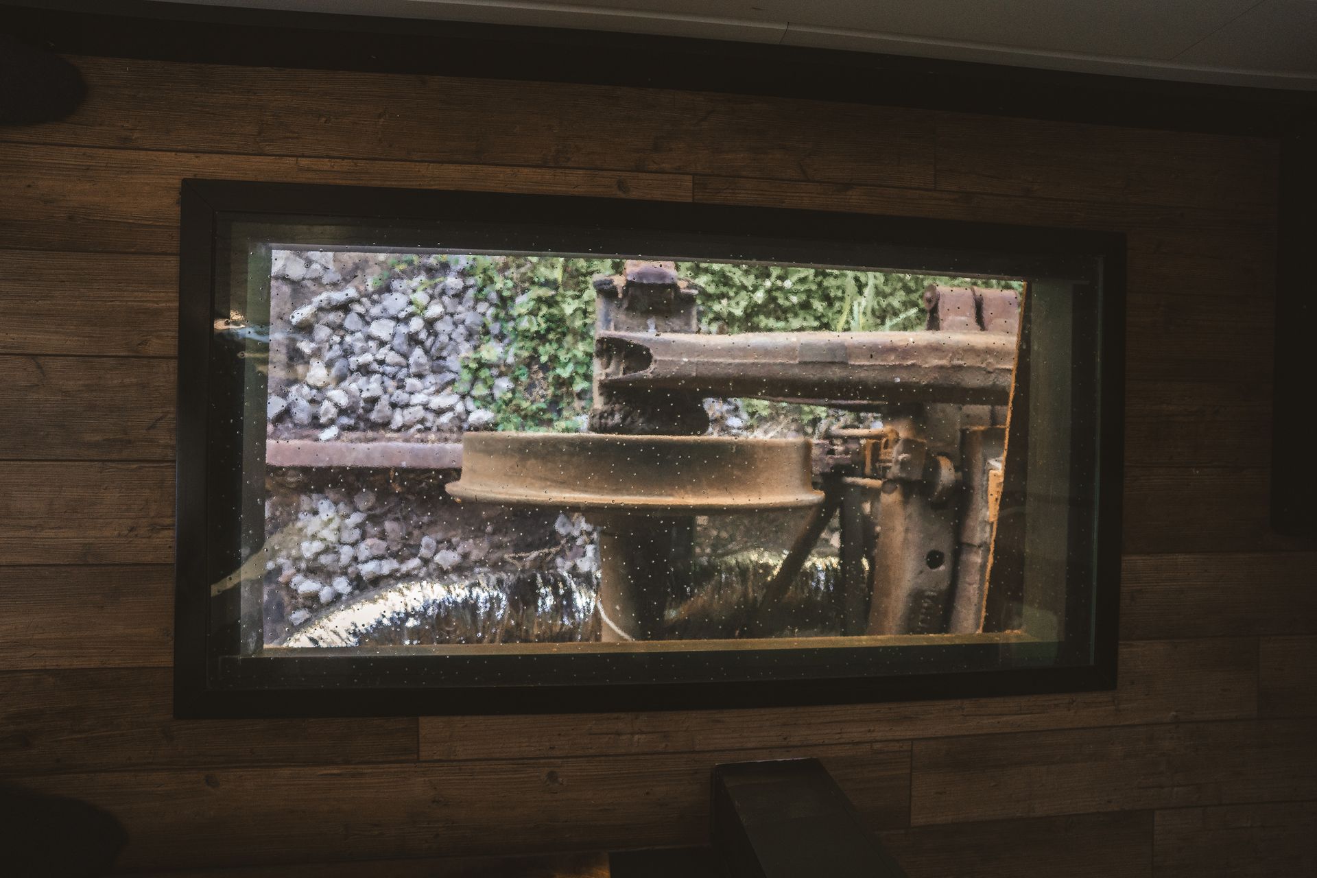 Window view of old, rusty machinery surrounded by gravel and green foliage.
